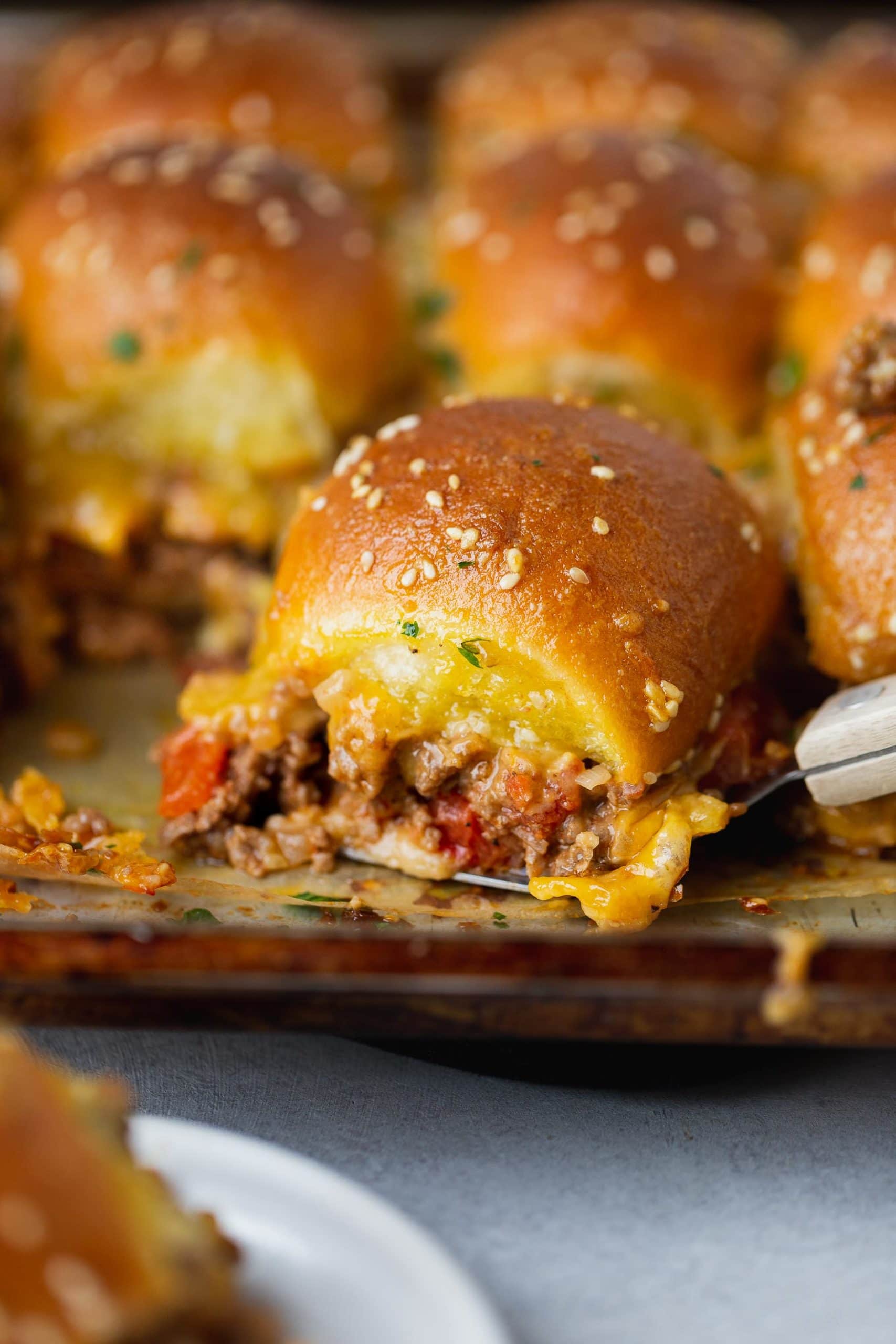 A cheeseburger slider filled with ground beef, melty cheese and chopped tomatoes. there are other sliders in the background.