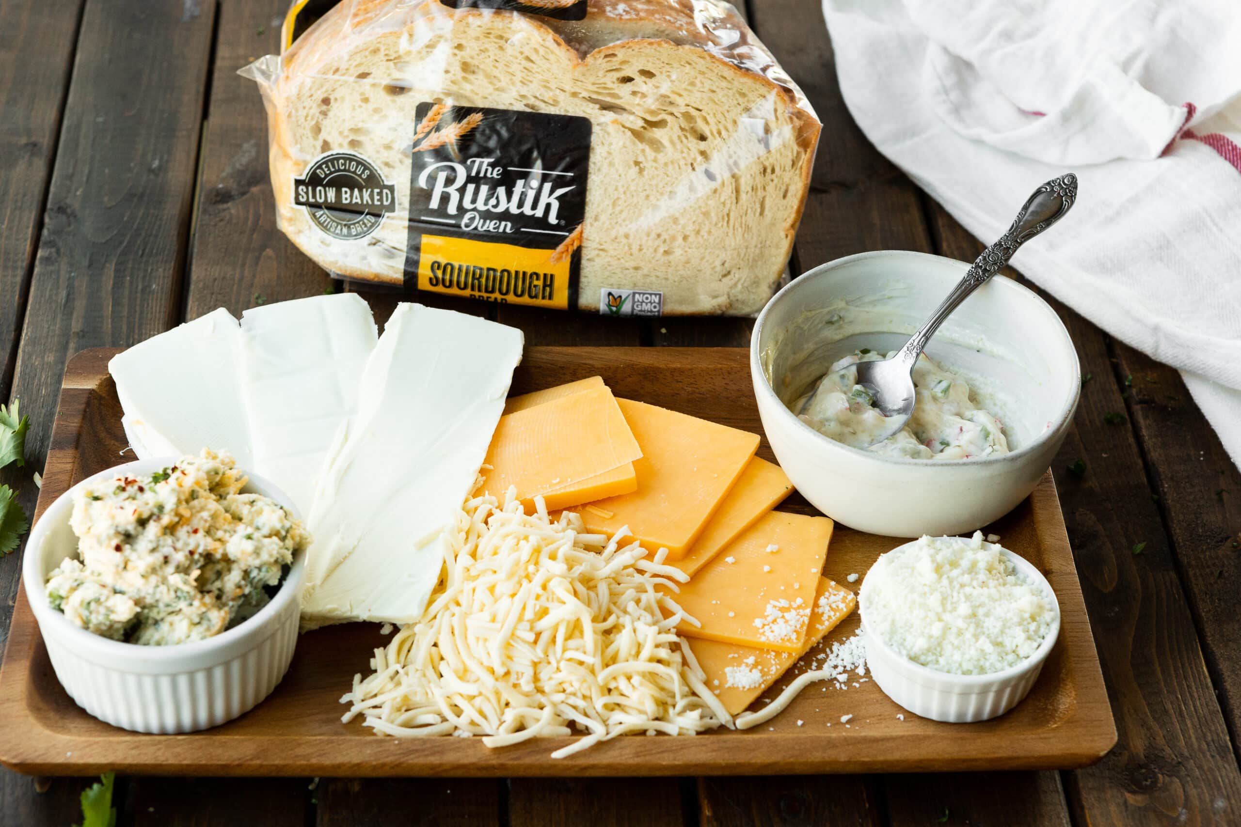 a photo of all the ingredients to make a Mexican grilled cheese sandwich sitting on a wooden cutting board including 4 different cheeses, a bag of Rustik sourdough bread, and a small bowl of the butter spread.