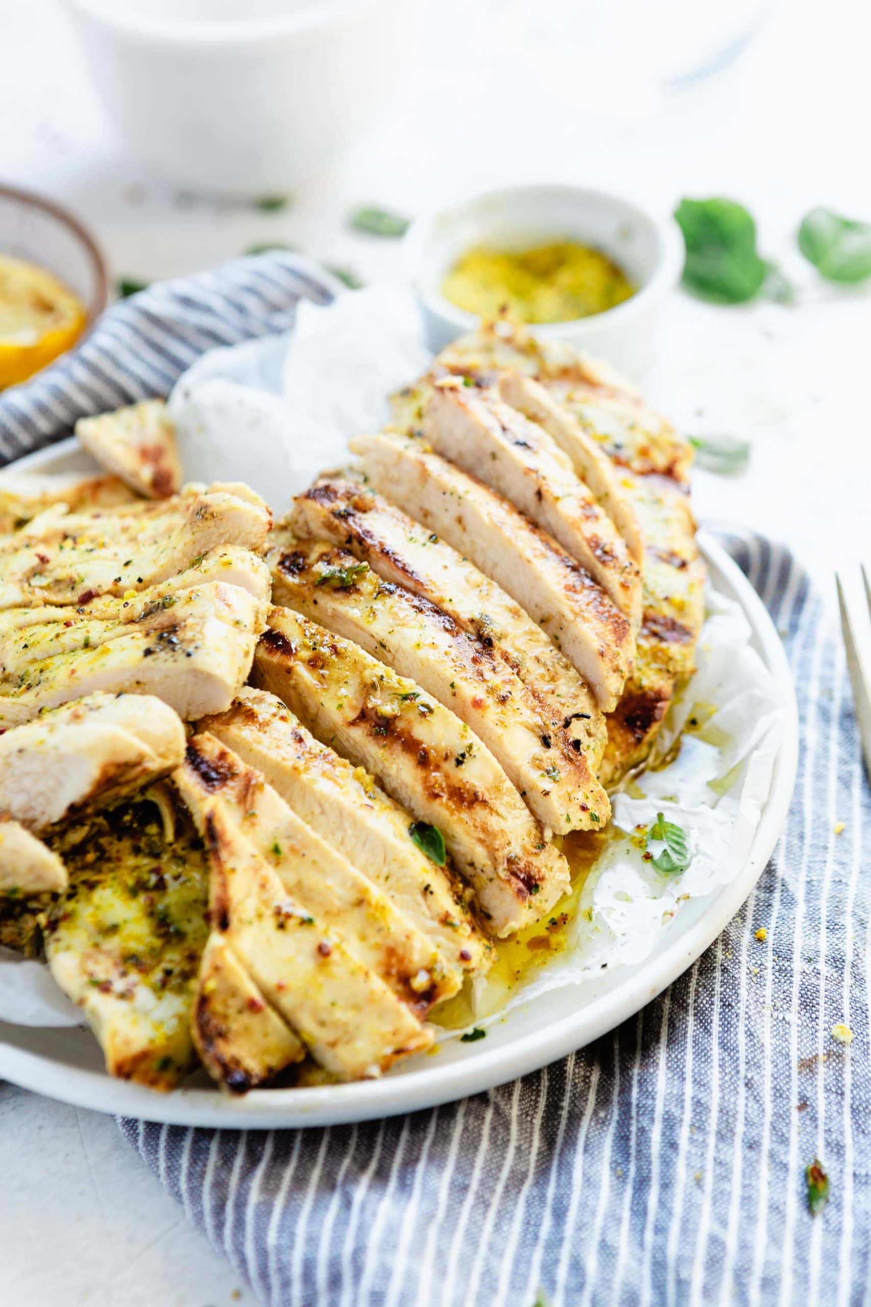 a plate of sliced, grilled chicken breasts on a blue and white striped cloth. Grilled lemon slices are in the background.