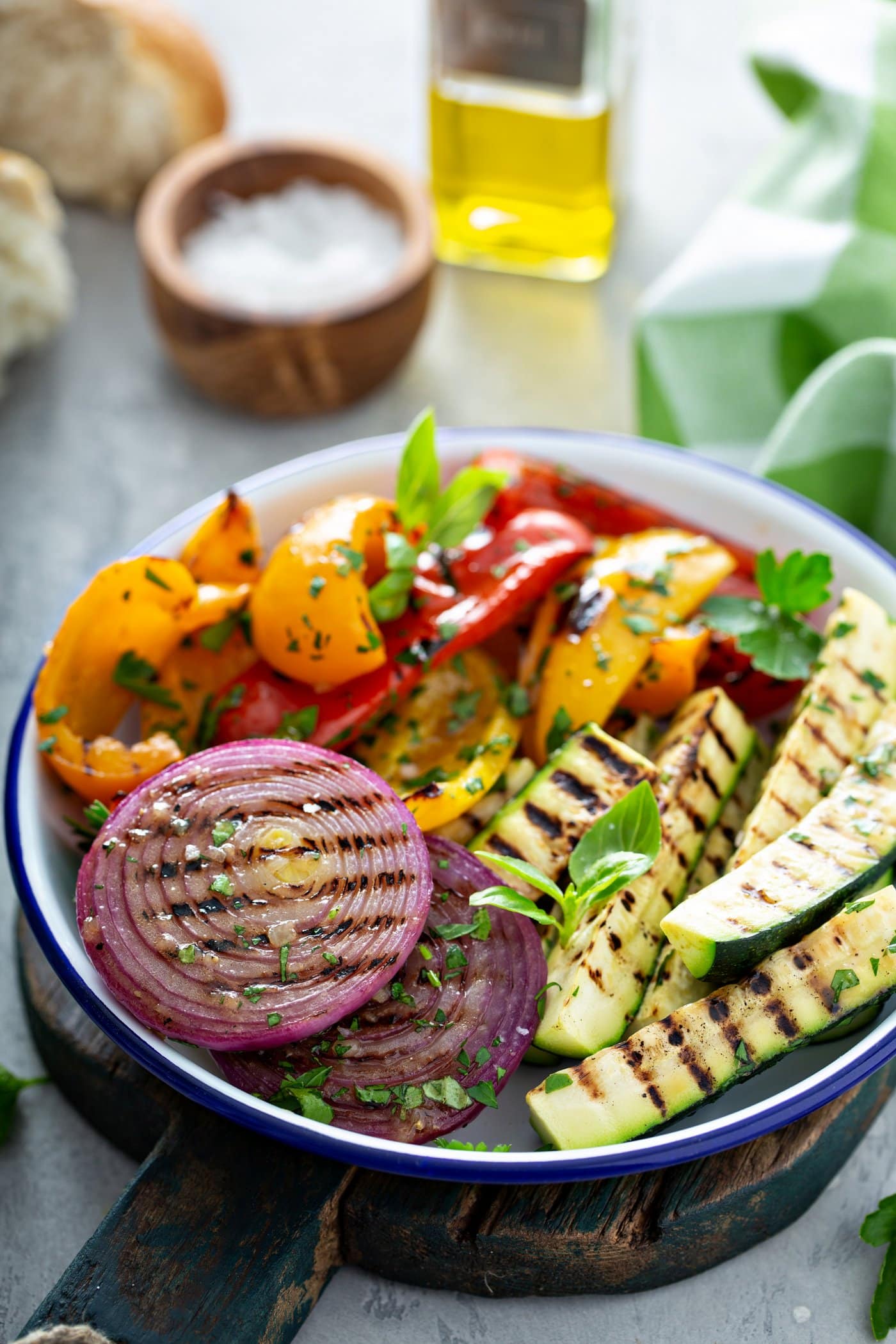 A serving dish filled with grilled veggies. There are sliced red onions, red and yellow bell peppers, and zucchini with grill marks. they are topped with a few parsley leaves. There is a wooden container of salt, a glass container of oil, and a green and white checked cloth in the background.