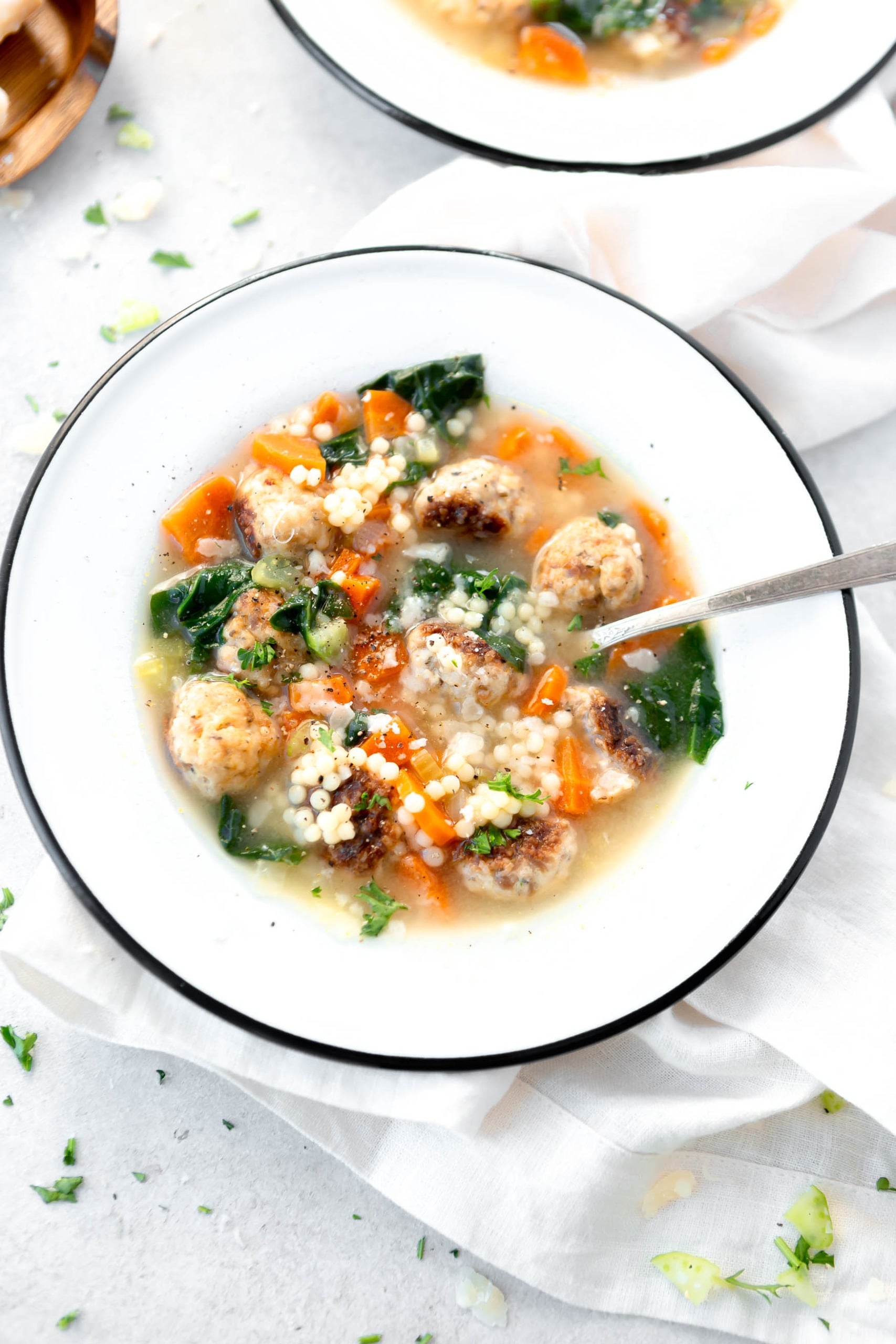 A bowl of Italian wedding soup. A spoon is in the soup and it is packed with meatballs, carrots and spinach.