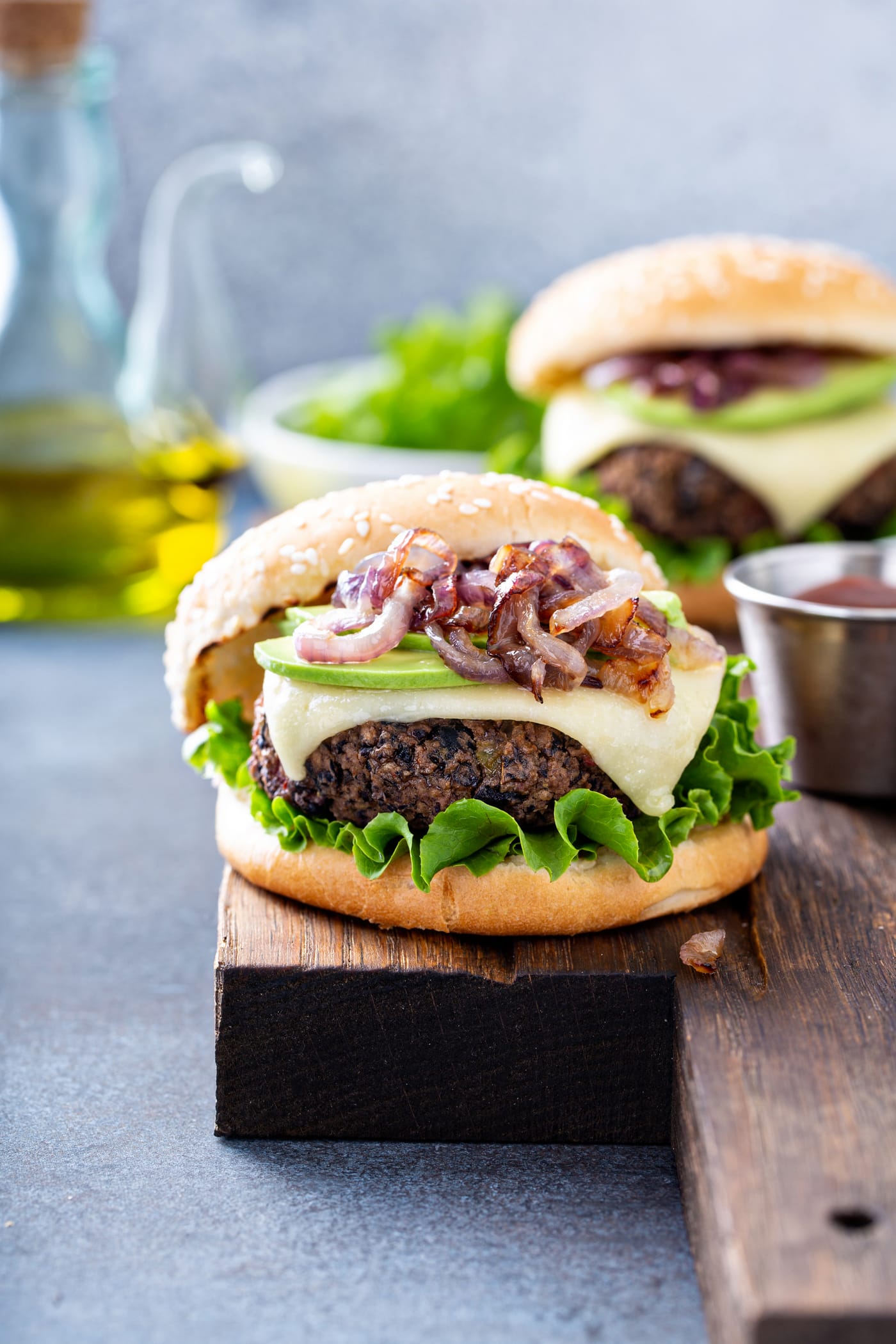 A table top with two black bean burgers on sesame seed buns. The burgers are topped with onions, avocados, cheese and lettuce. There is a bowl of lettuce and a container of oil in the background.