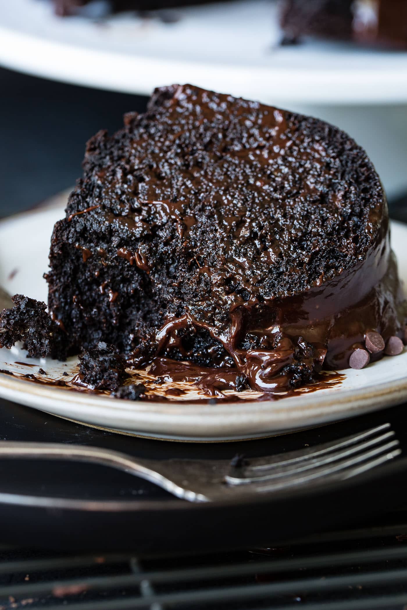 A slice of zucchini chocolate bundt cake on a white dessert plate. The cake is dark, moist chocolate with a chocolate glaze. A fork is next to the plate.