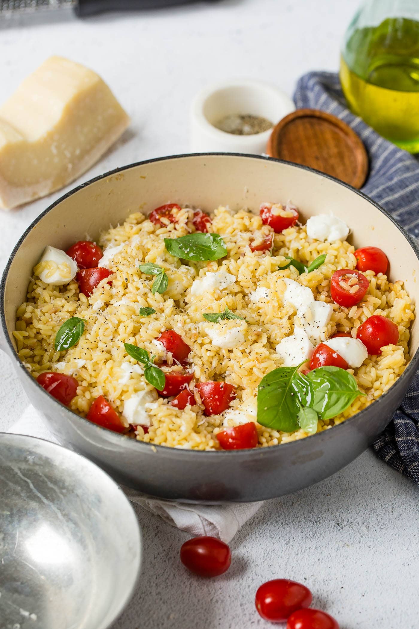 A serving bowl of orzo caprese salad. It is loaded with tomatoes, fresh basil and mozzarella cheese. Parmesan cheese, salt, and tomatoes are next to the bowl of orzo.