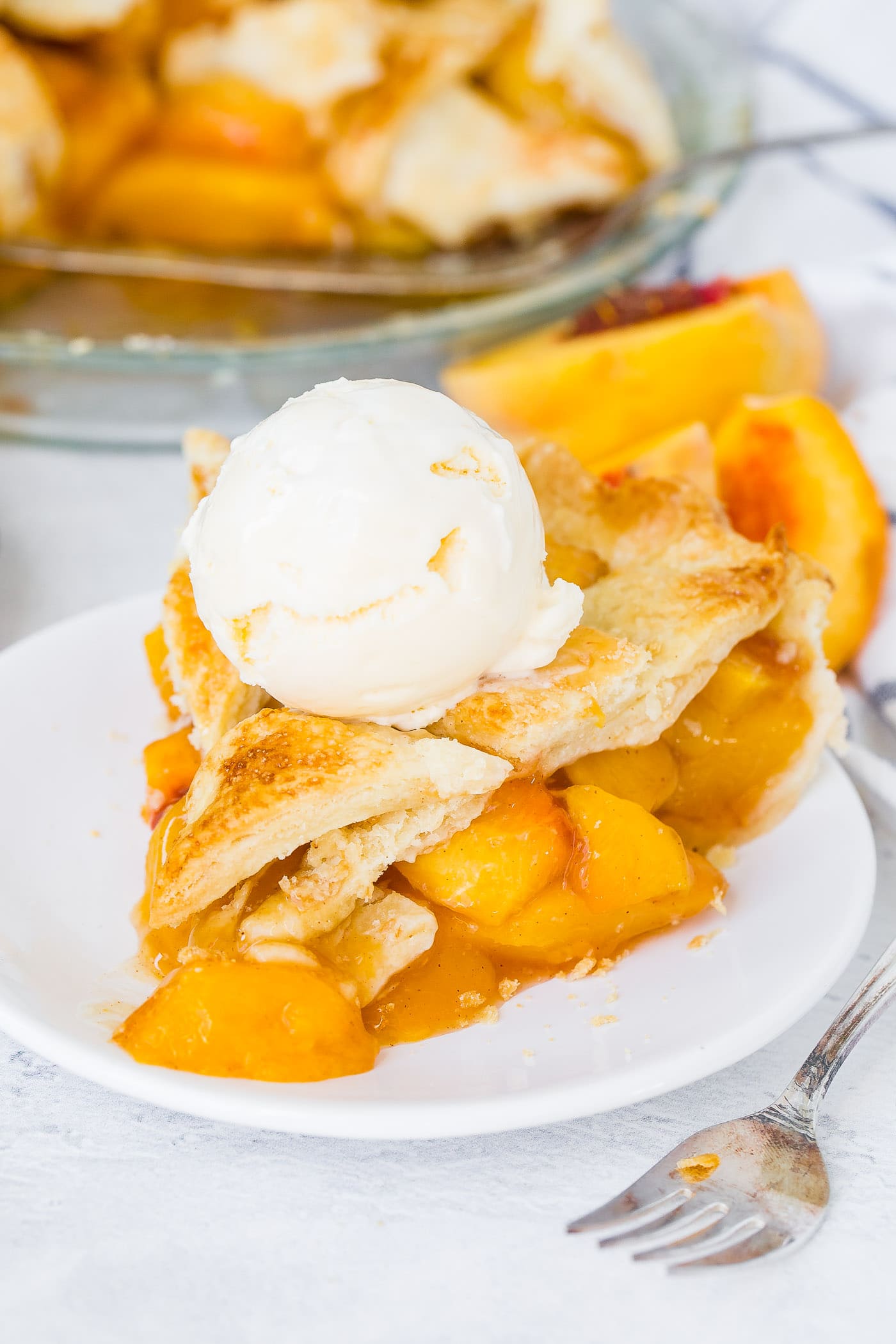 A photo of a slice of fresh peach pie with lattice crust on a white plate with a scoop of vanilla ice cream on top.