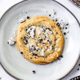 A dessert plate holding a large cookies and cream cookie. The cookie is loaded with bits of cookies and cream candy bar and there are bits of candy bar on the plate.