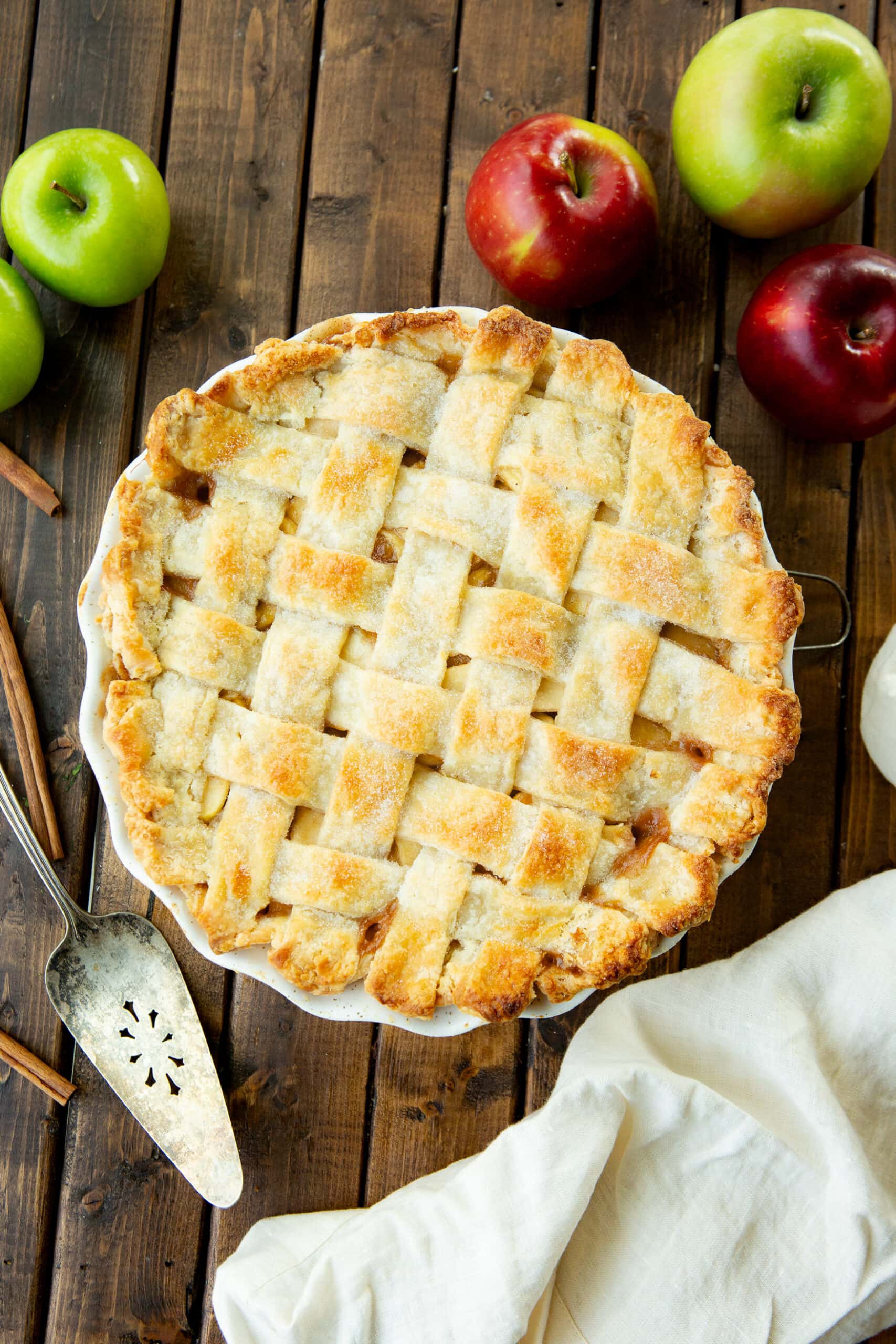 a photo of a whole baked apple pie with a lattice top sitting on a wooden table with a silver pie server sitting next to it and red and green apples scattered around it.