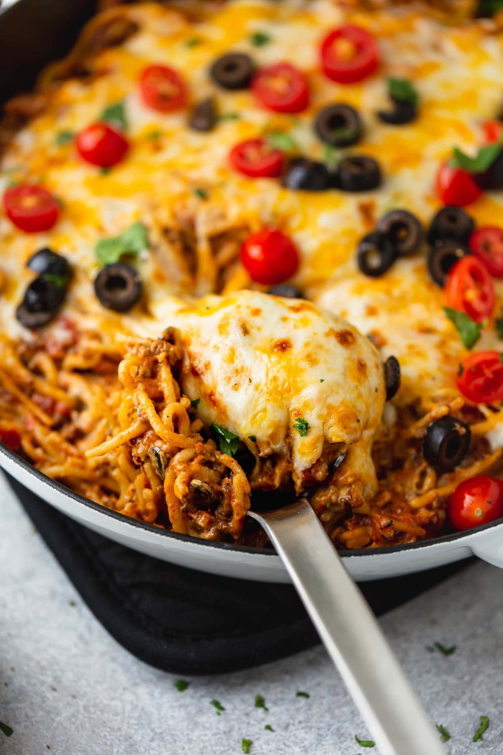 A skillet full of cheesy ground beef taco spaghetti. A serving spoon is in the pot and the spaghetti is topped with melted cheese, tomatoes, olives, green onions and cilantro.