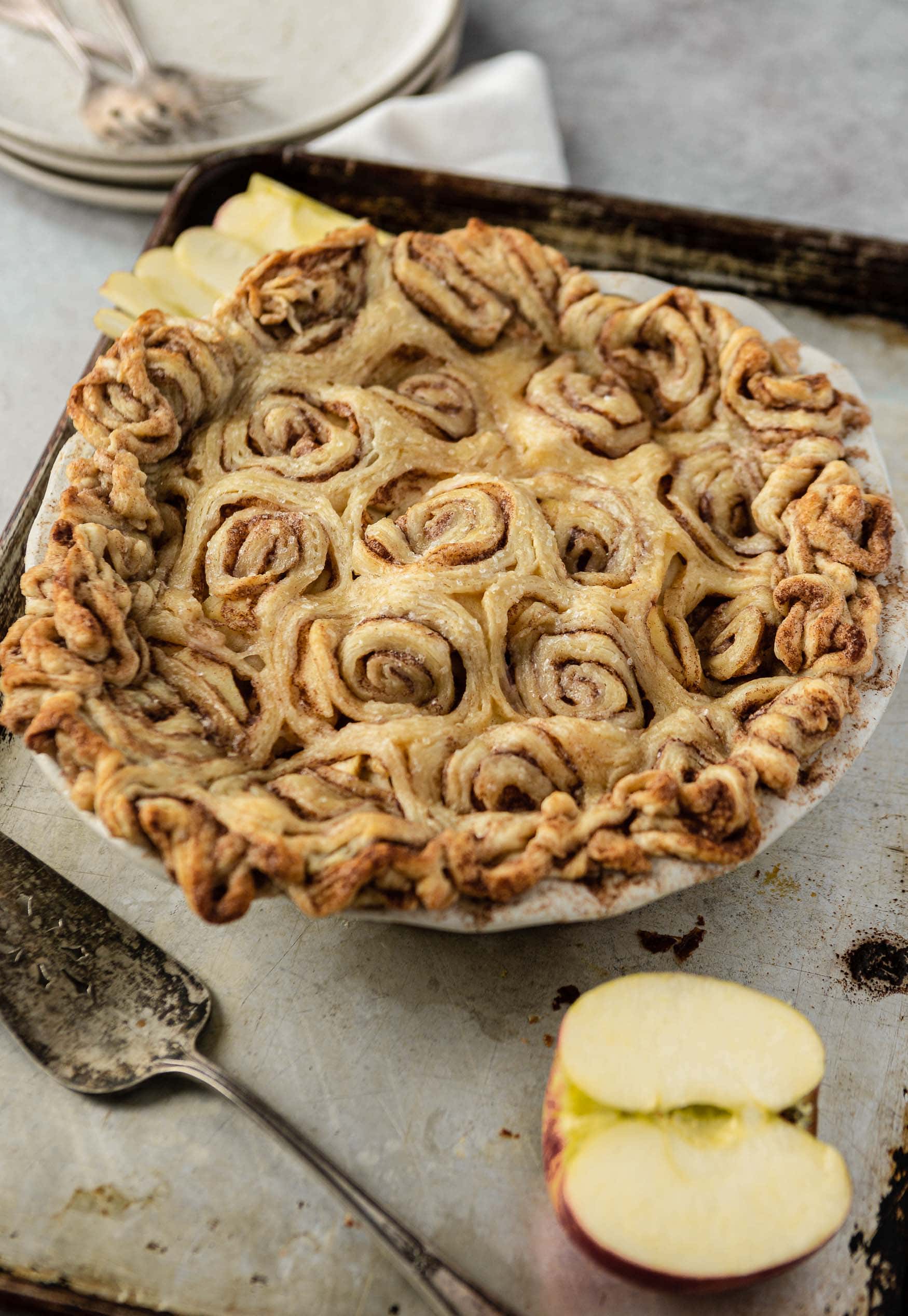 A baked cinnamon roll apple pie. The top crust is cinnamon rolls. There is a sliced apple and a pie server next to the pie.