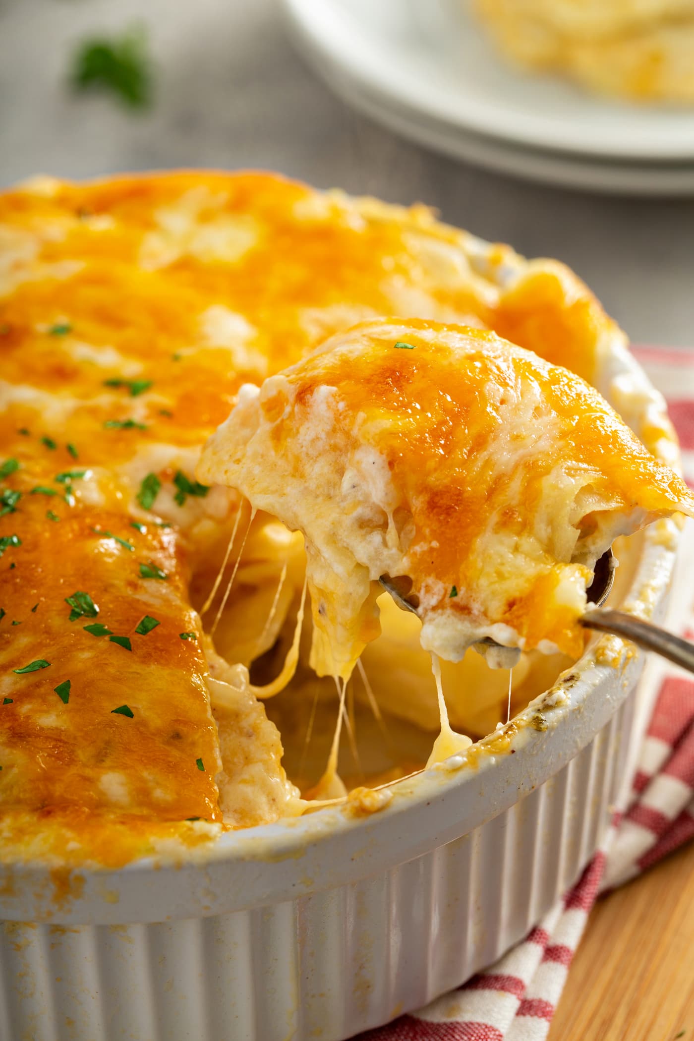 A casserole dish filled with cheesy potatoes. The cheese is melted to perfection and there are strings of cheesy hanging from the serving spoon. 