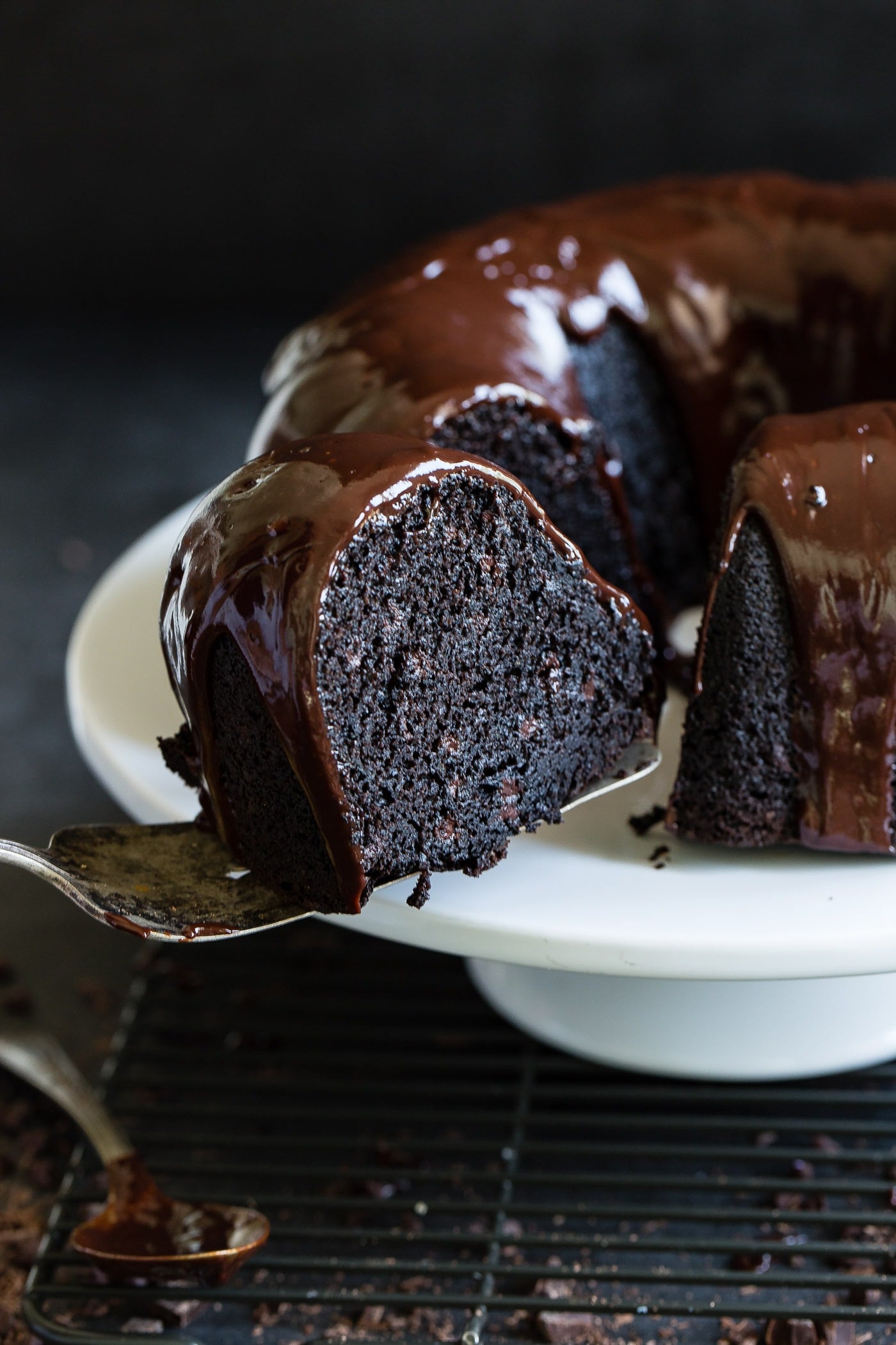A slice of zucchini chocolate bundt cake being removed from the cake. The inside of the cake is dark, moist chocolate and there is a chocolate glaze on top of the cake.