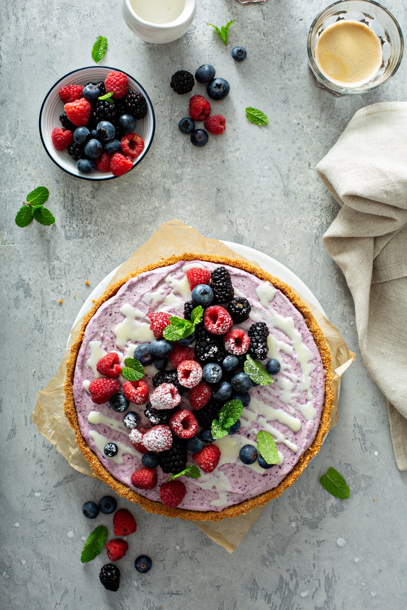 A whole white chocolate berry pie with mixed berries on top. There are mint leaves sprinkled on top of the berries and on the table top. A bowl of mixed berries and a napkin are in the background.