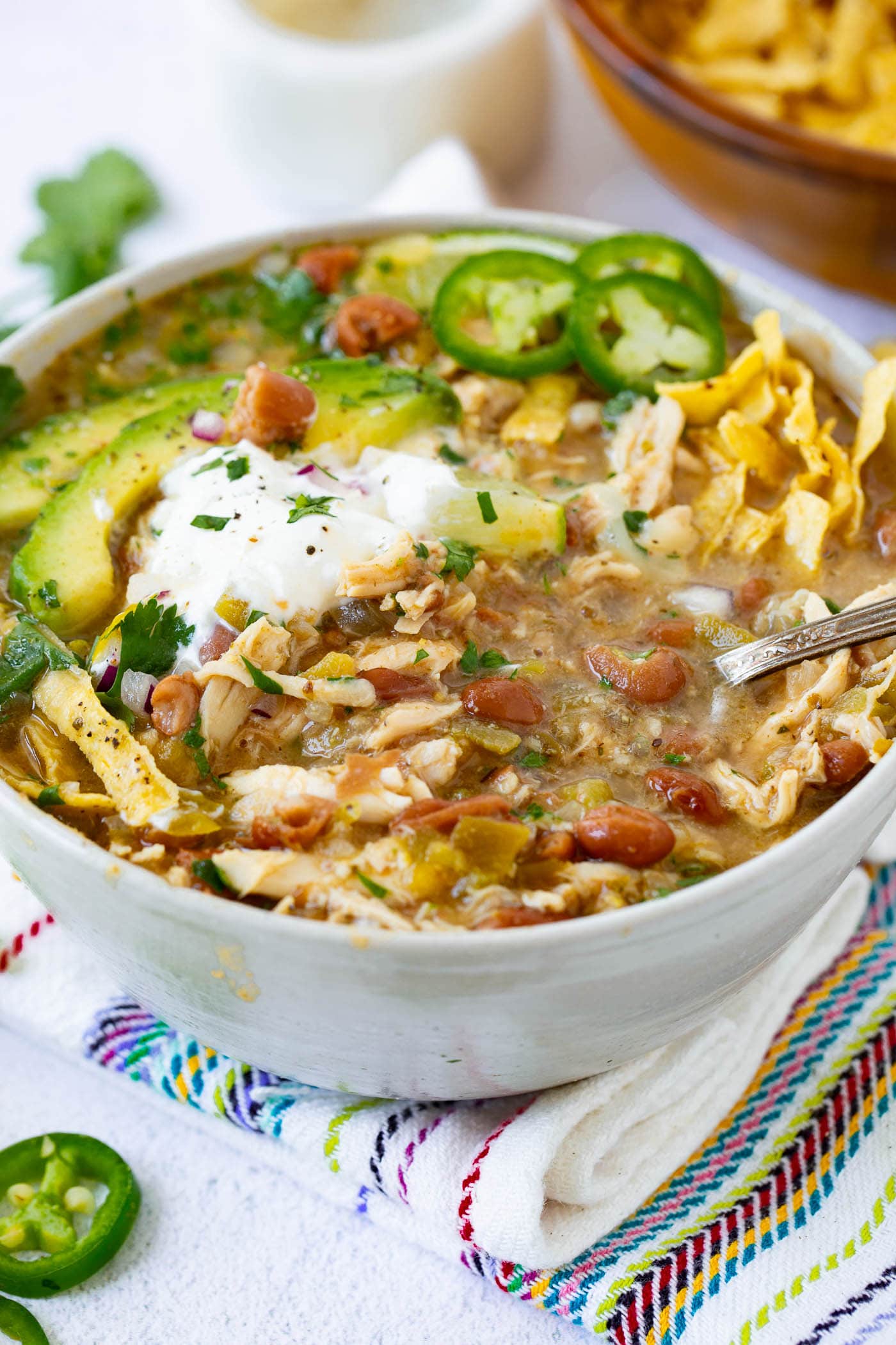 a photo of a bowl full of white chicken chili topped with a dollop of sour cream, avocado slices, tortilla chip strips and slices of fresh jalapeno with a spoon in the bowl