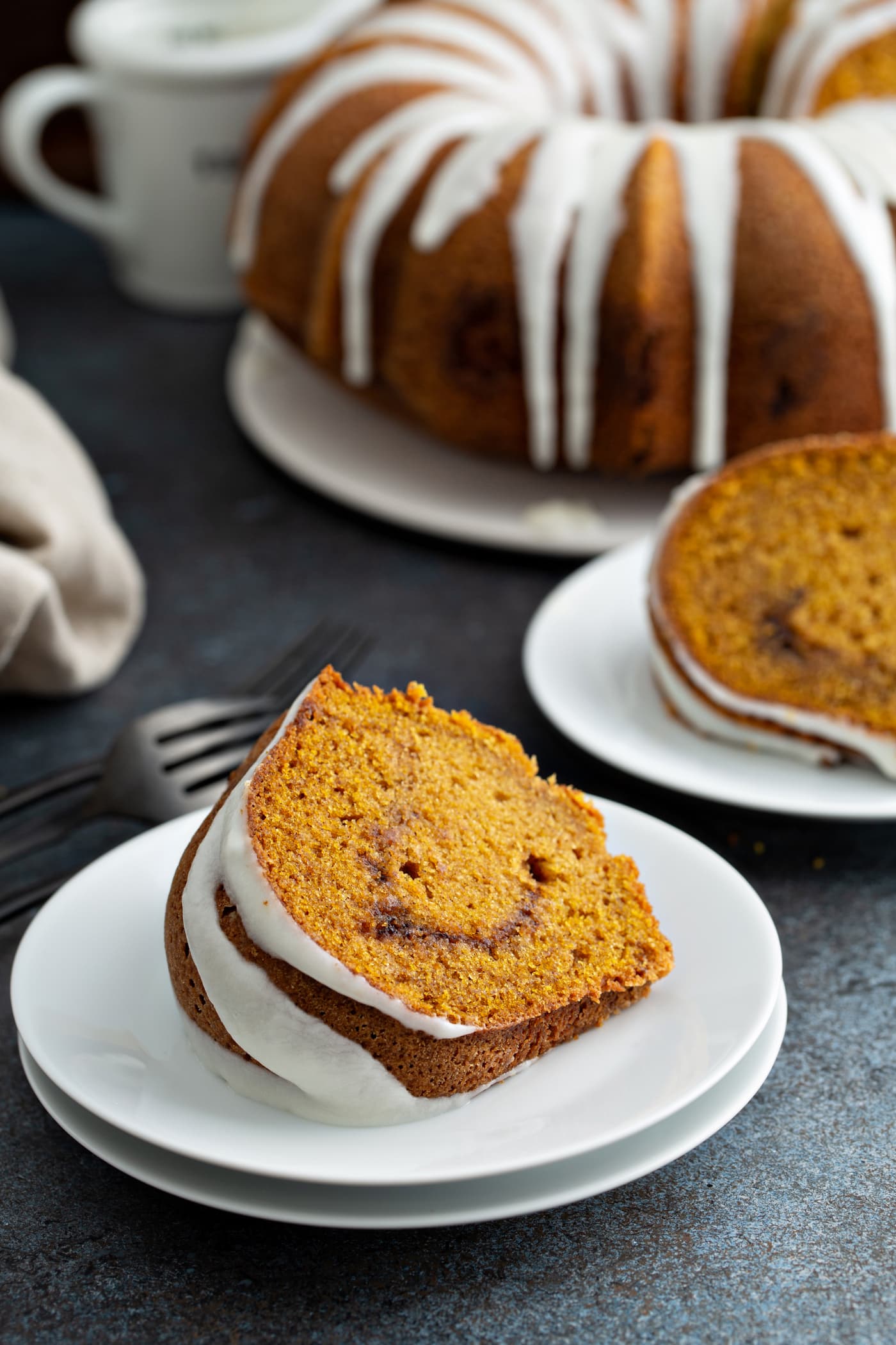 Two slices of sour cream cinnamon swirl pumpkin bundt cake on dessert plates. The cinnamon swirl center is visible and the cake top is drizzled with white glaze. The rest of the bundt cake with glaze drizzled over the top is in the background. A fork and napkin are next to one of the dessert plates.
