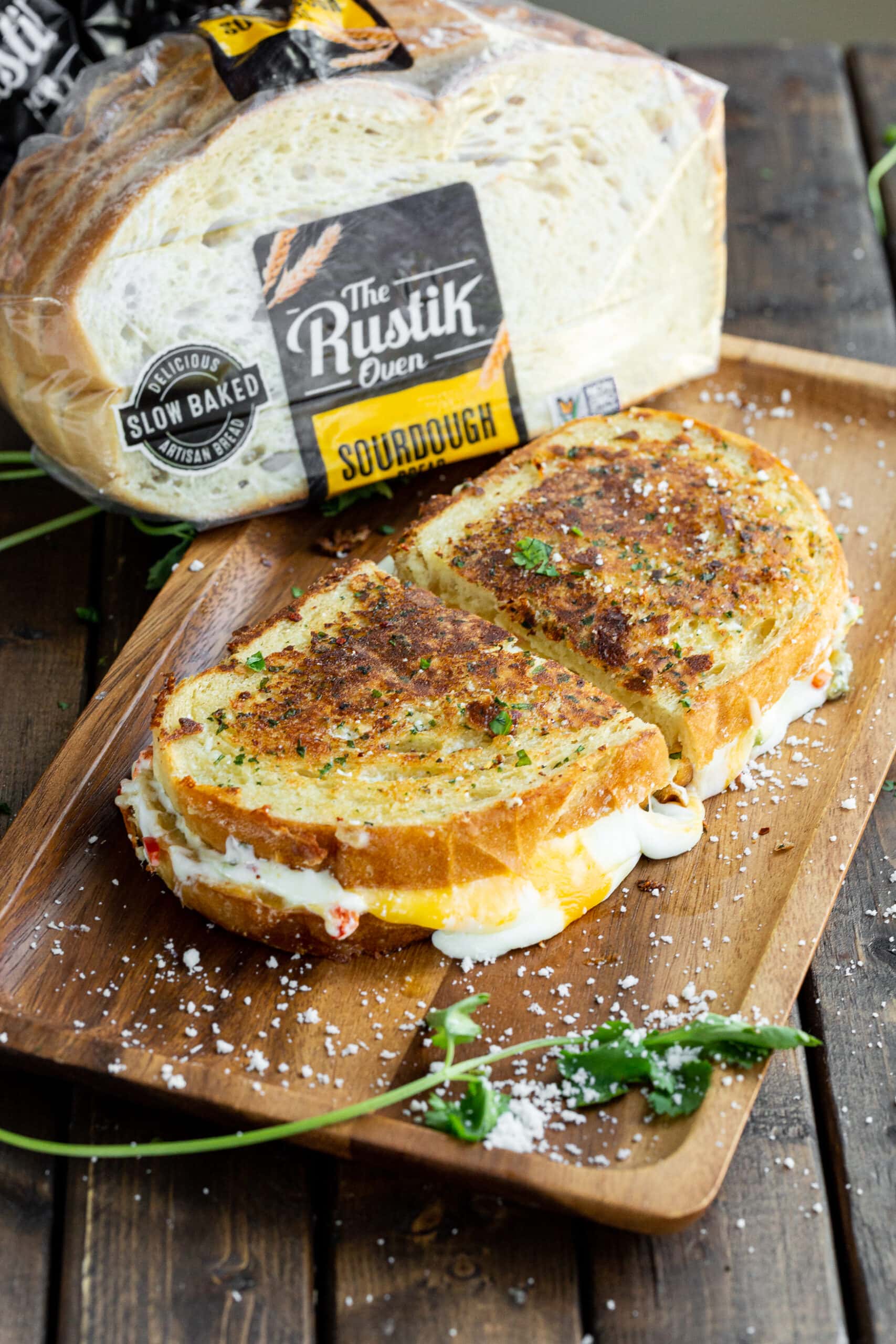 a photo of a perfectly golden grilled cheese sandwich with melted orange and white cheeses oozing out the sides of the sandwich which has been cut in half. There is a bag of The Rustik Oven sourdough bread sitting behind the sandwich.