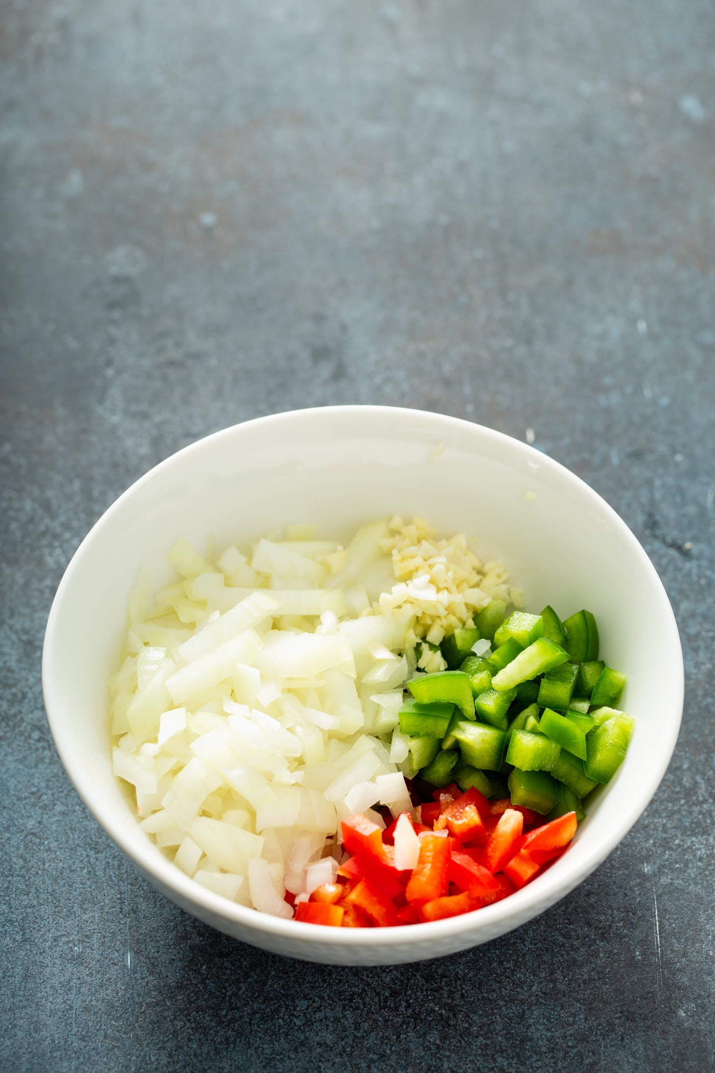A white bowl with chopped onions, and chopped red and green peppers.