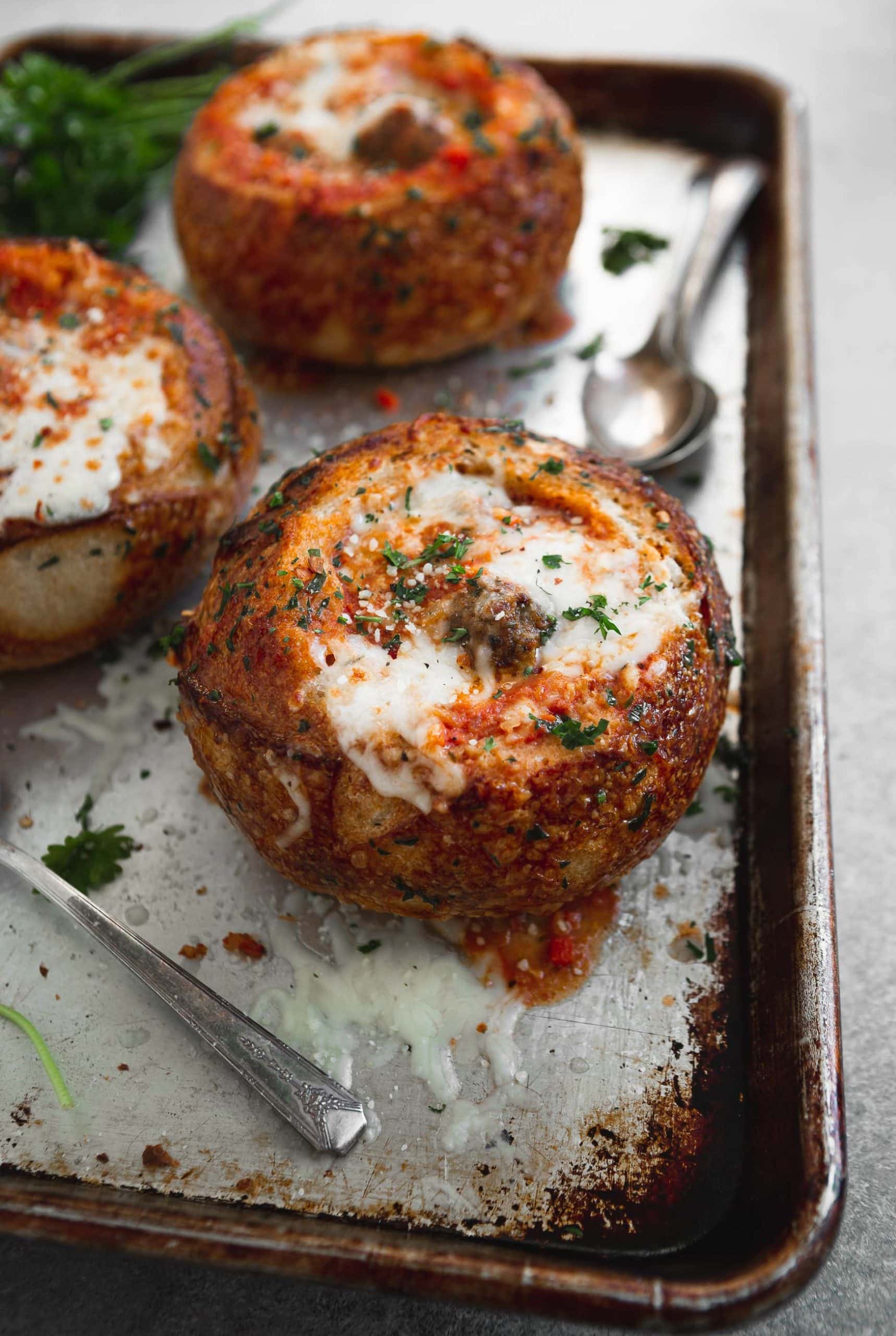 Three toasted bread bowls filled with meatball parmesan soup. there is melted cheese sprinkled with fresh parsley on top.