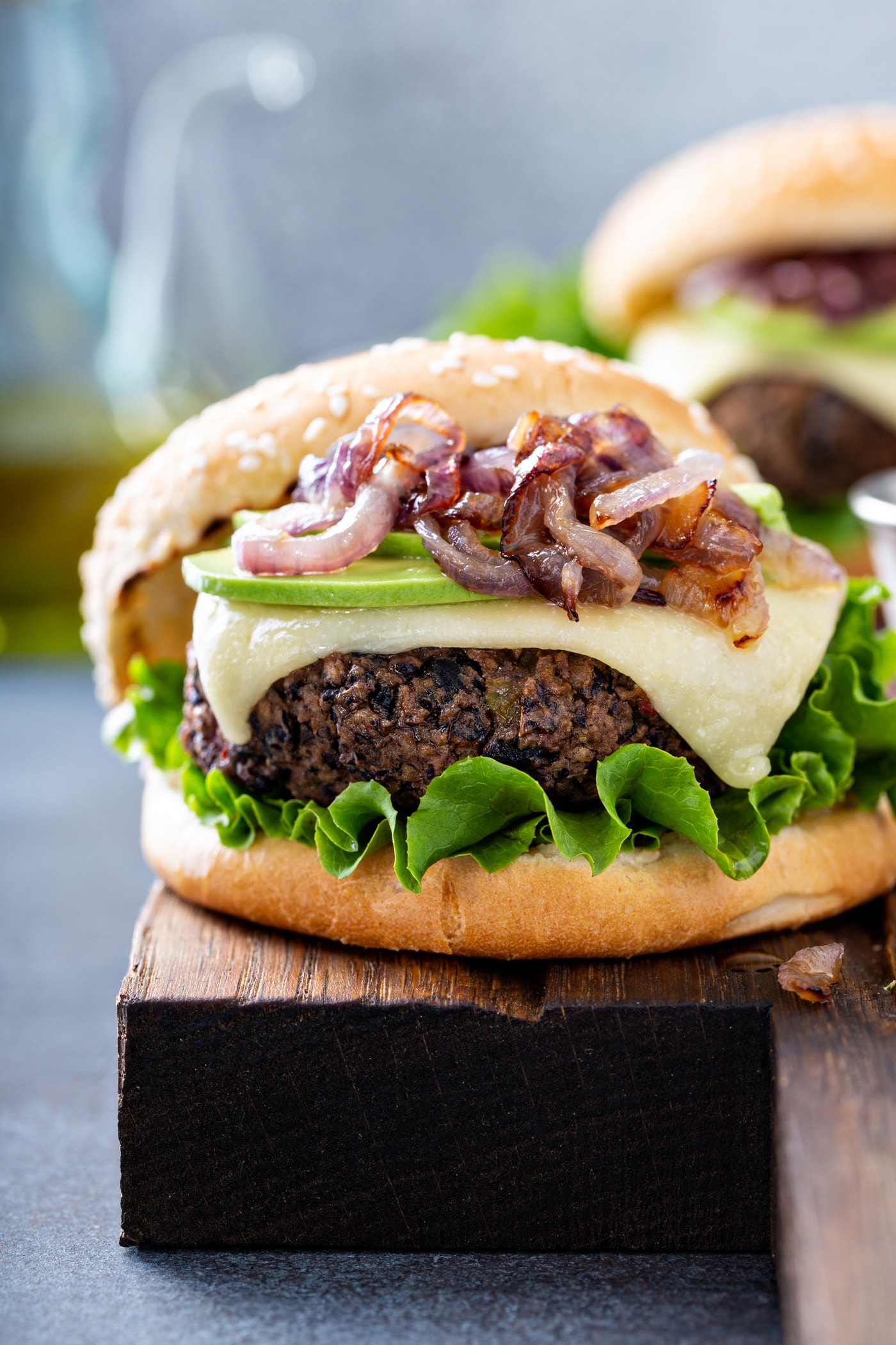 A close-up of a black bean burger on a sesame seed bun, topped with onions, avocado, cheese and lettuce.