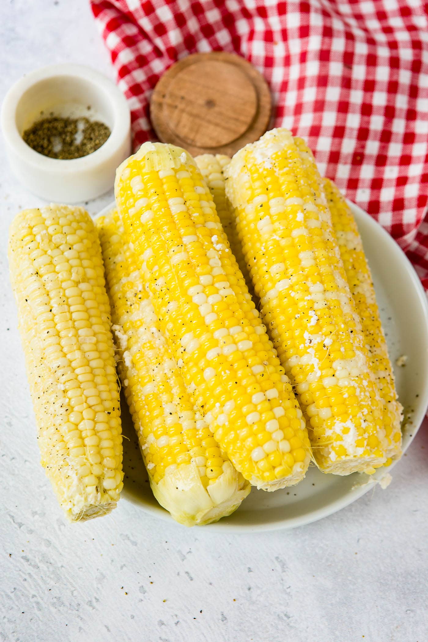 A serving plate with 5 ears of corn. The plate of corn is sitting on a red and white checked tea towel and there is a small container of 