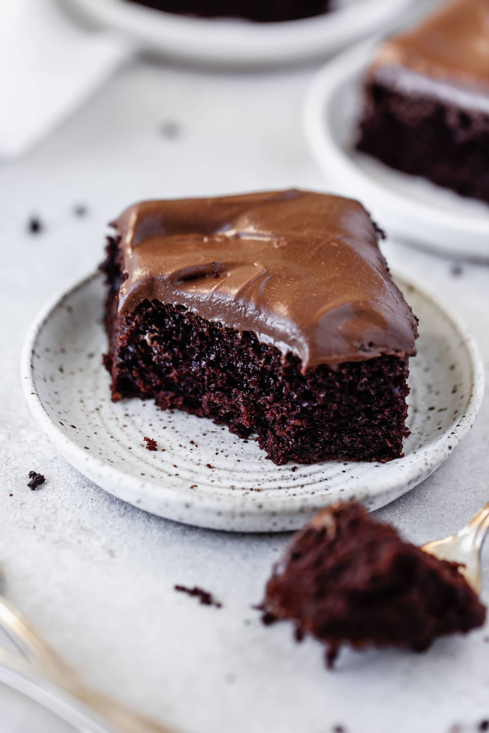 A piece of chocolate mayonnaise cake that has a bite taken out of it. It is frosted with chocolate cream cheese frosting. Other servings of cake are in the background.