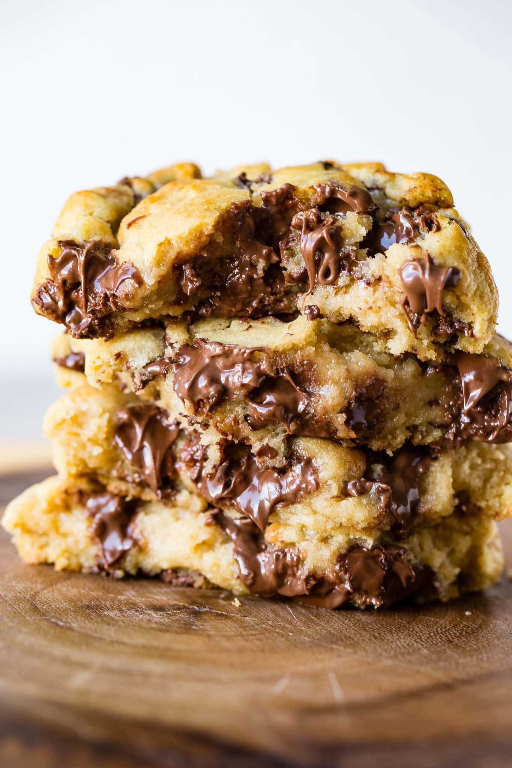 a photo of four thick melty chocolate chip cookies that have been broken in half stacked on top of each other on top of a wooden plate.