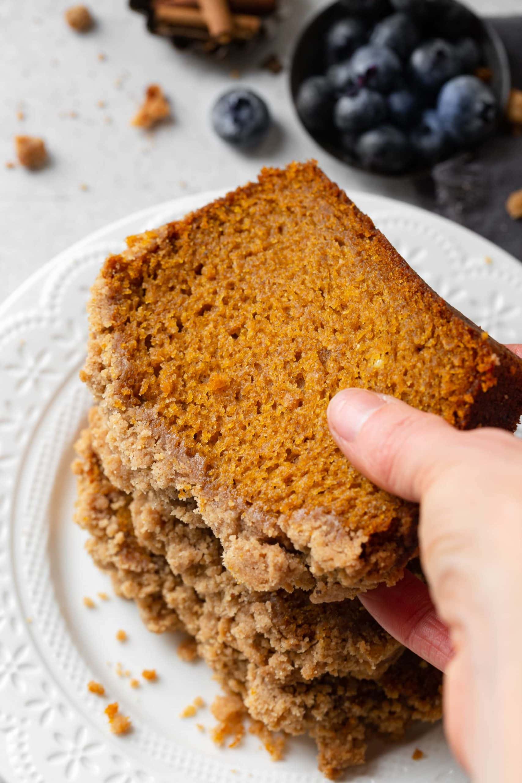 a photo of someone picking up a slice of pumpkin bread with a thick streusel topping from a pile of slices of bread.