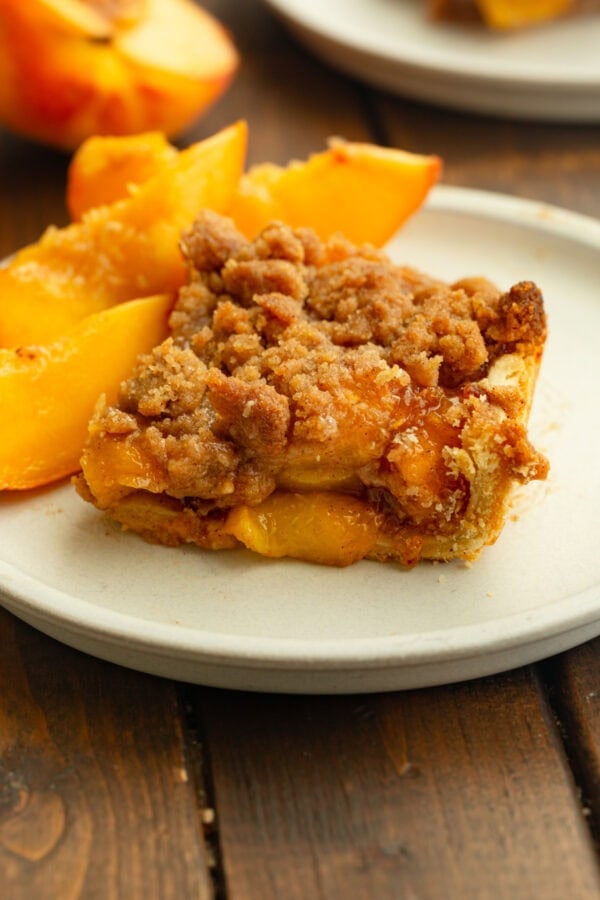 A slice of peach crumble bar on a white plate, topped with golden-brown streusel and served with fresh peach slices.