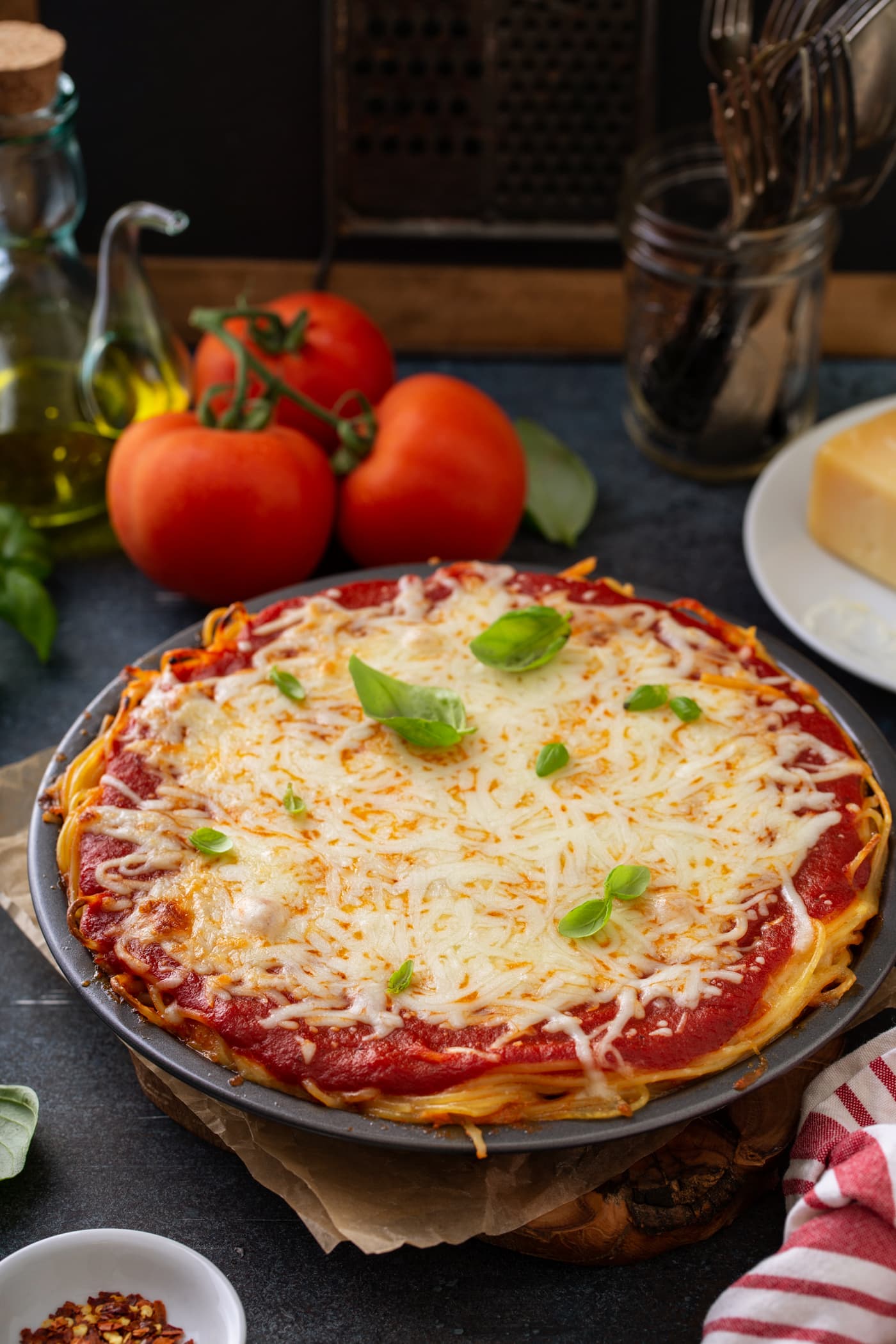 A pie dish filled with baked spaghetti pie. There are layers of spaghetti, spaghetti sauce and melted cheeses. and a few basil leaves on top. Three tomatoes, olive oil and bay leaves are in the background.