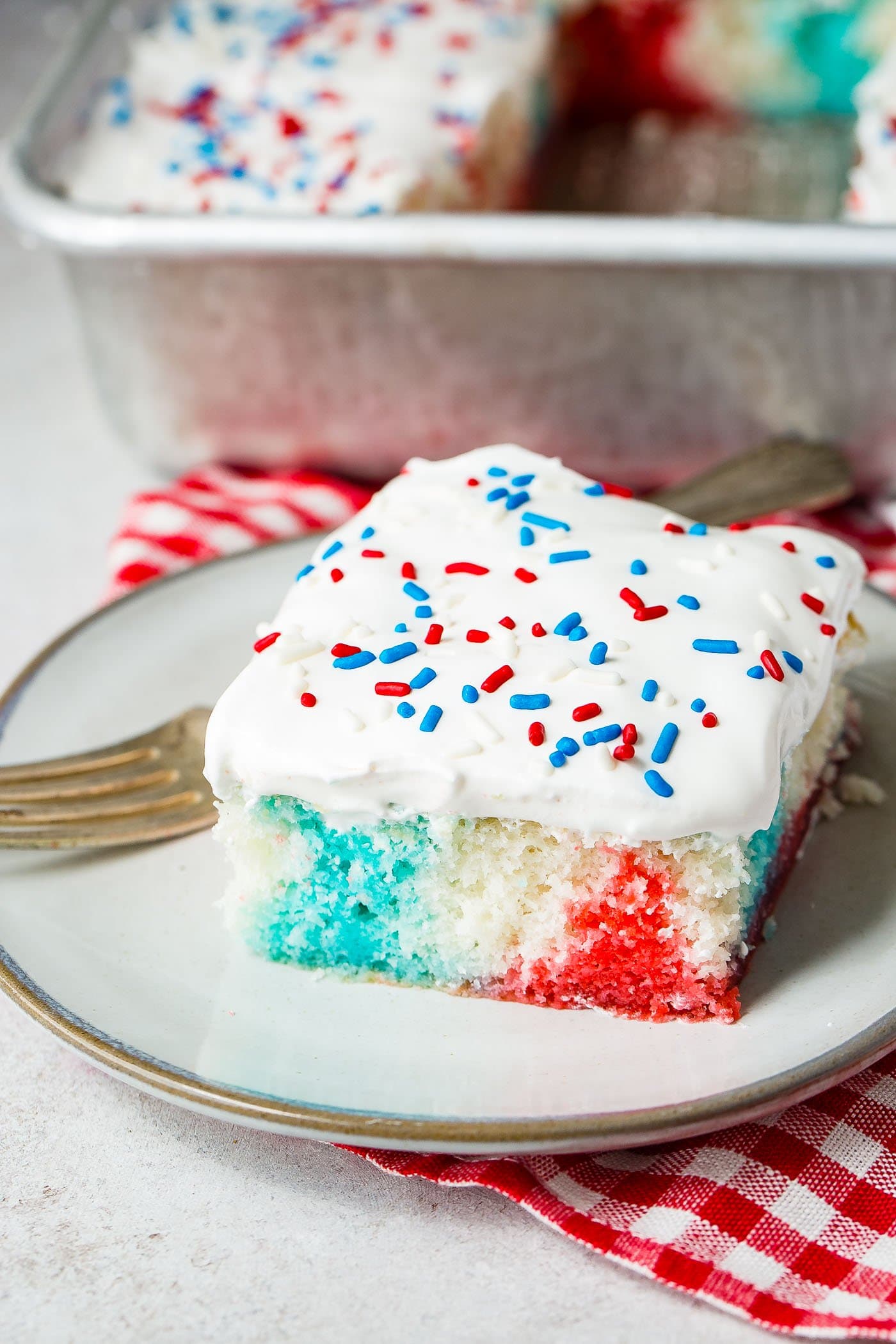 A piece of red, white and blue poke cake on a white dessert plate with a fork resting next to the cake. There is a red and white checkered napkin in the background.
