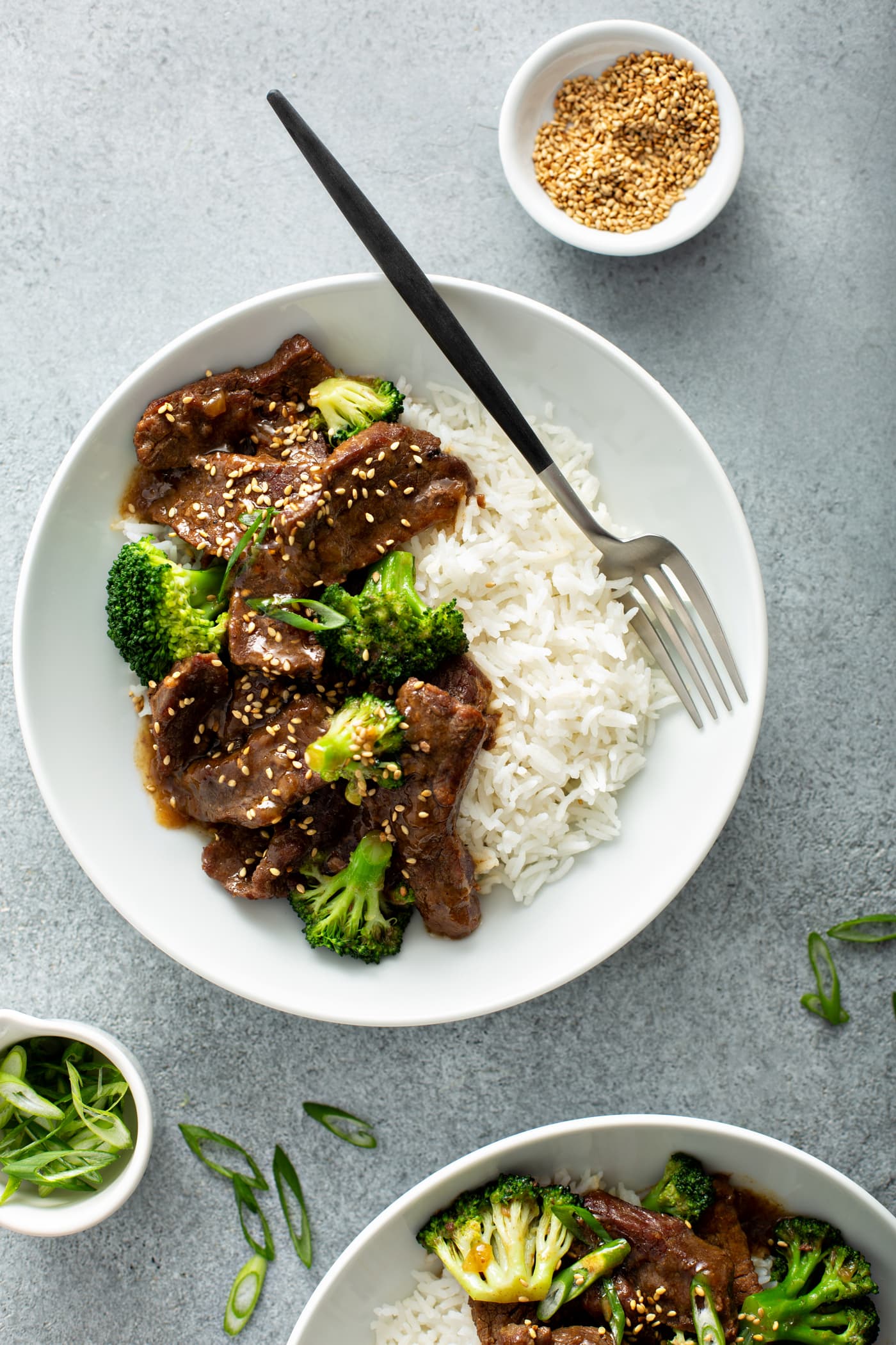 A photo of a white bowl with a serving of beef and broccoli in it along side some white rice. There is also a small dish of toasted sesame seeds next to the bowl.