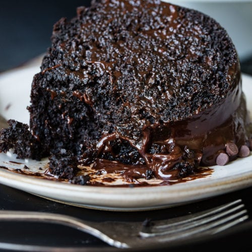 A slice of zucchini chocolate bundt cake on a white dessert plate. The cake is dark, moist chocolate with a chocolate glaze. A fork is next to the plate.