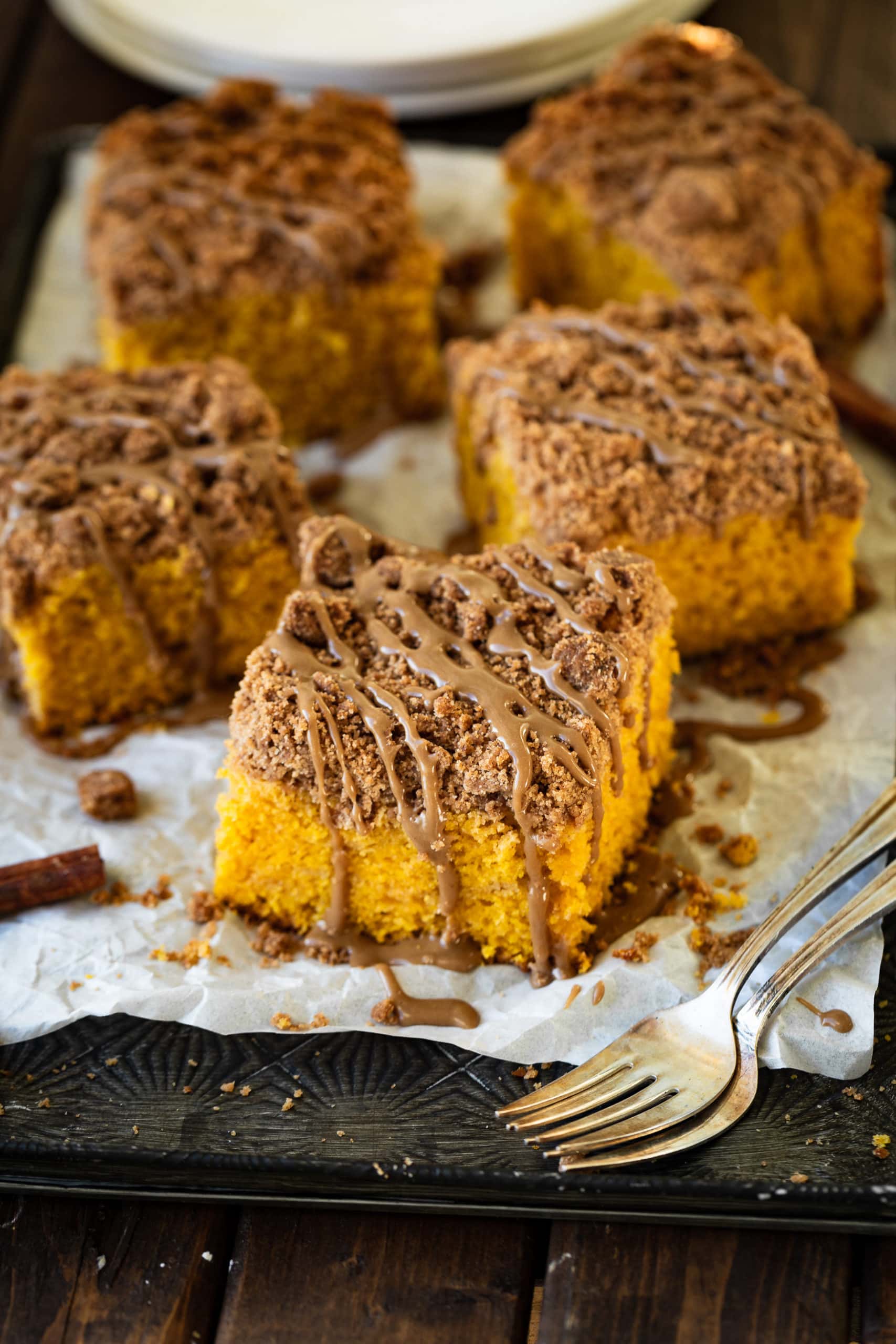 a photo of several pieces of pumpkin coffee cake sitting on a piece of parchment paper on a baking sheet. The coffee cake pieces are topped with cinnamon streusel and a thick maple glaze.