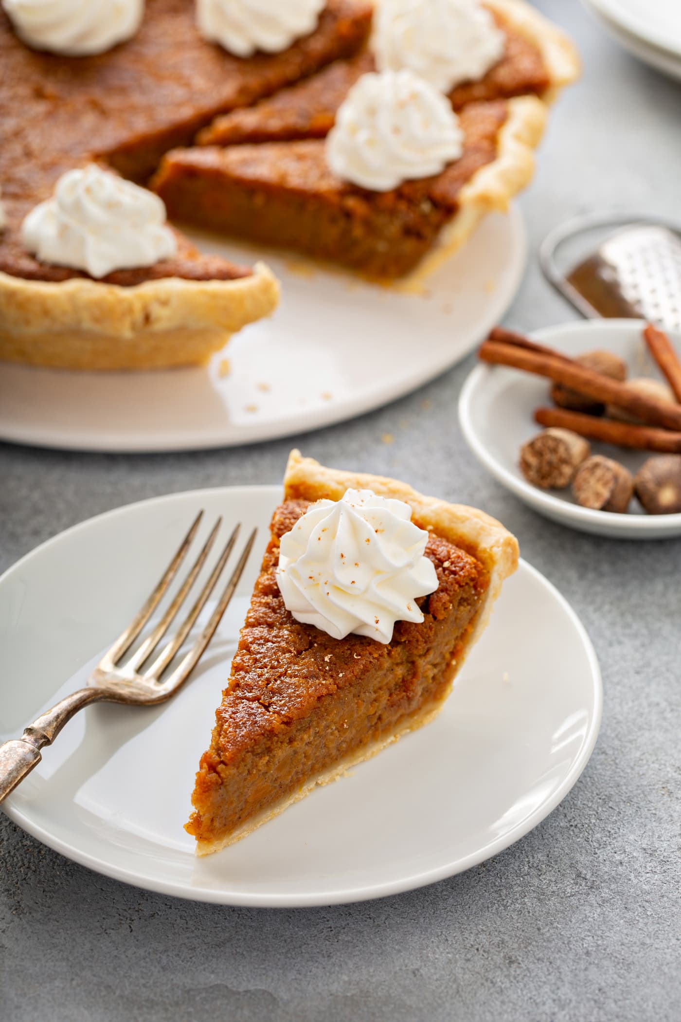 A photo of a slice of sweet potato pie sitting on a white plate with the whole sweet potato pie in the background.