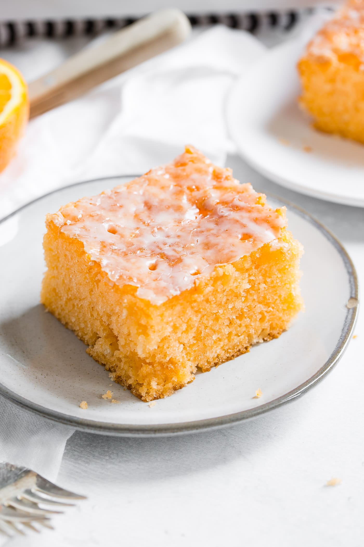 A photo of a piece of orange jello cake on a small white plate with another serving in the background and a fork in the foreground.