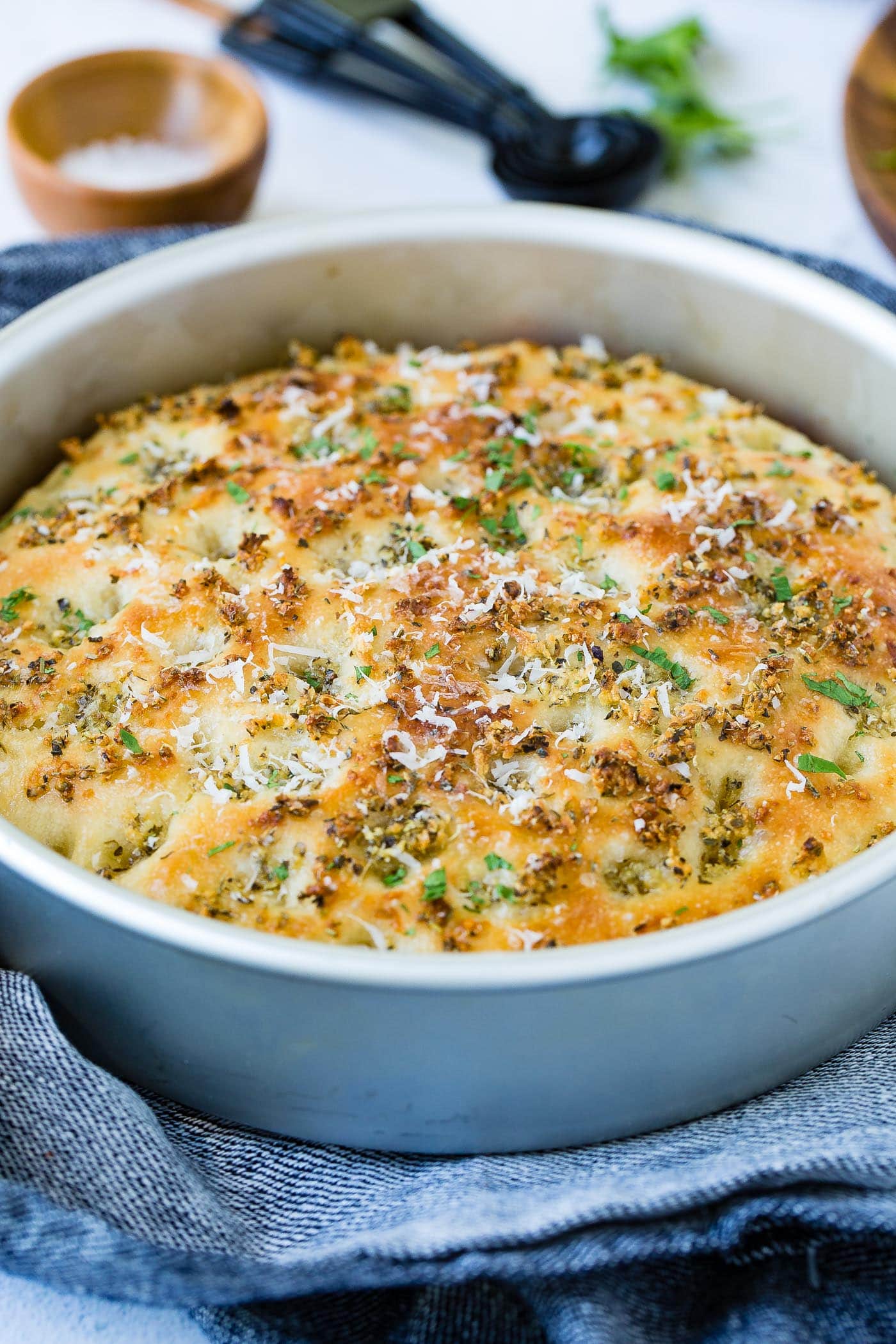 A round baking dish with baked garlic parmesan focaccia bread. the bread is golden brown and has bits of green herbs in it. There are fresh herbs, measuring spoons and a small container of salt in the background.