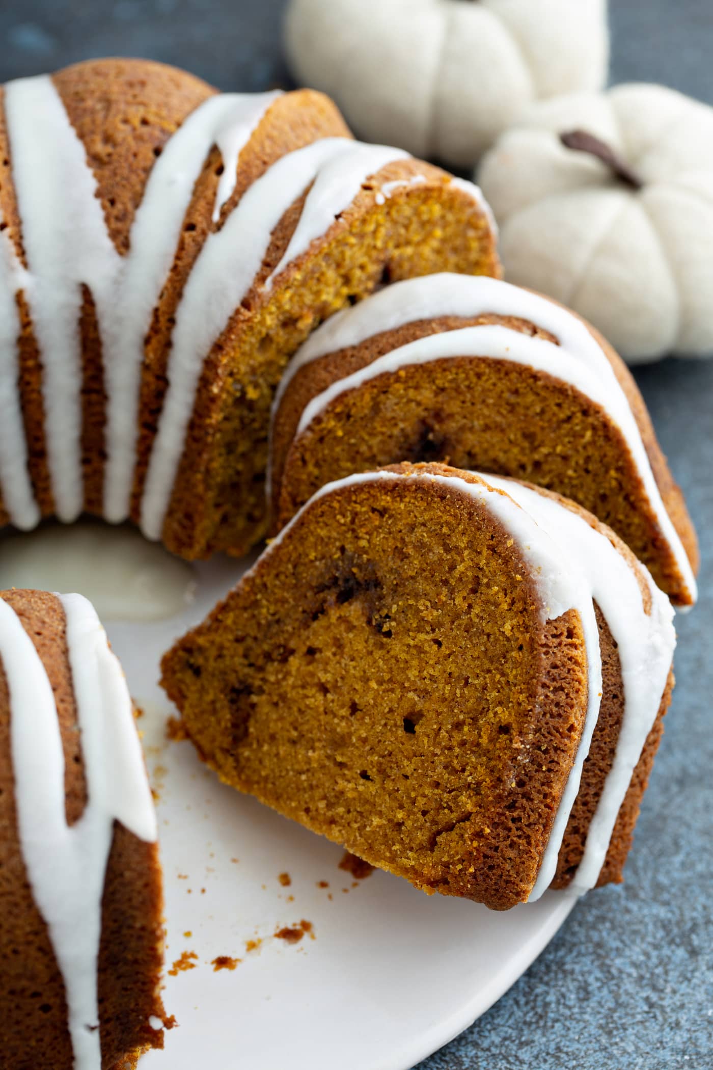 A sour cream cinnamon swirl pumpkin bundt cake on a cake platter. A couple of pieces of cake have been sliced. The cake has a white drizzled frosting on top.