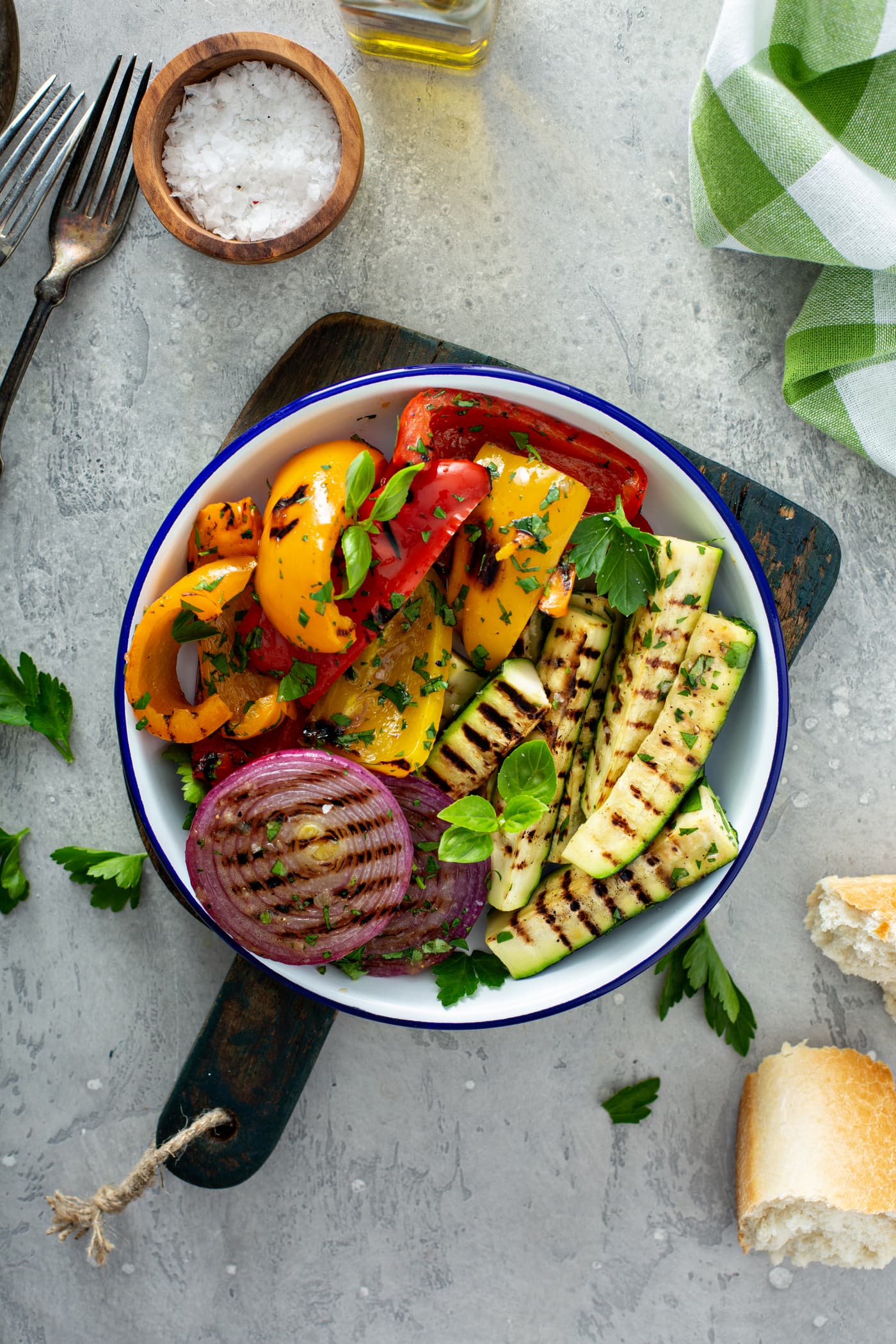 A serving plate of grilled vegetables sitting on a wooden cutting board. The grilled red onion, red and yellow bell peppers, and zucchini are topped with parsley leaves. A few parsley leaves are scattered on the table 