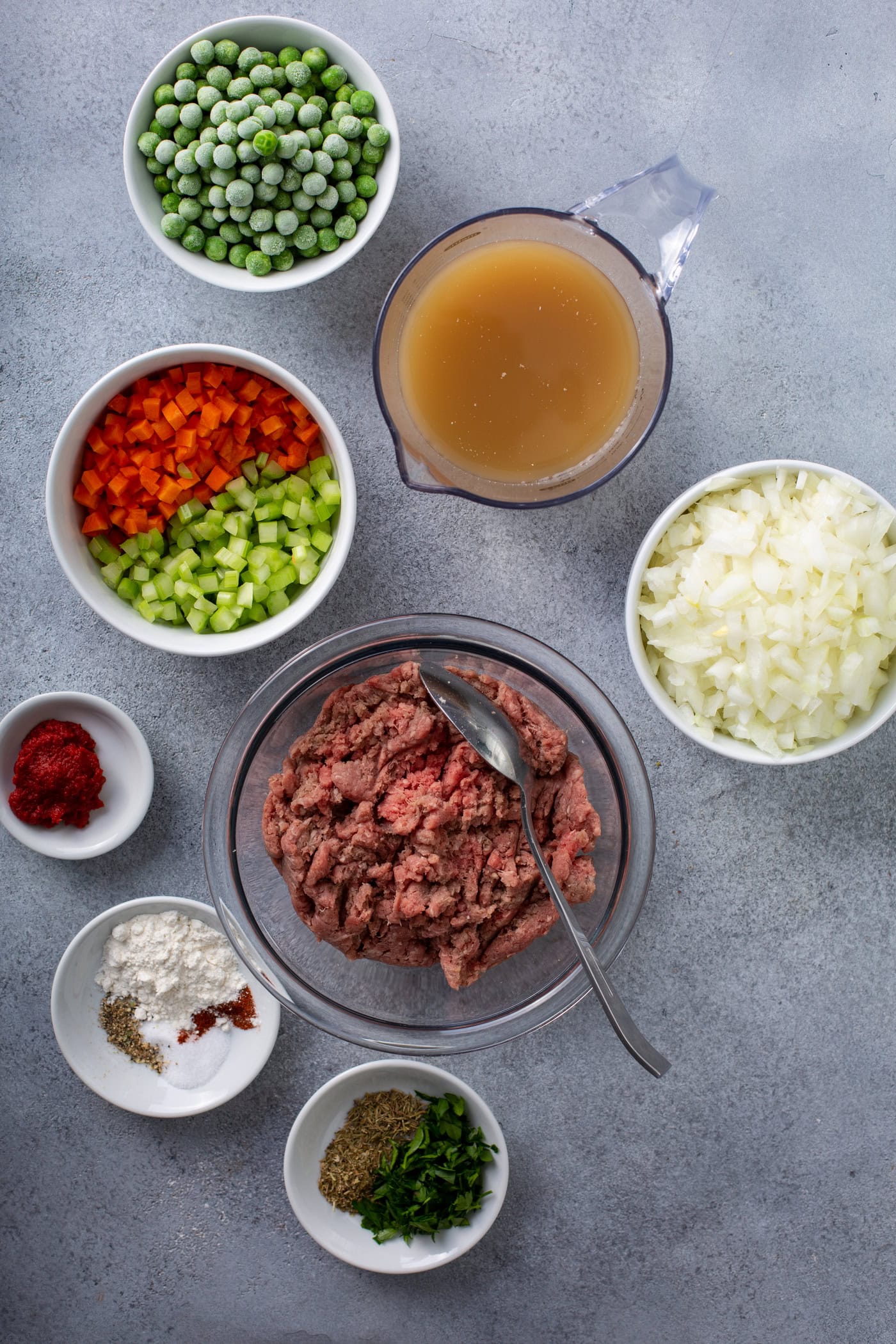 A table top with containers of ground beef, onions, chopped red and green peppers, spices, peas, and broth.