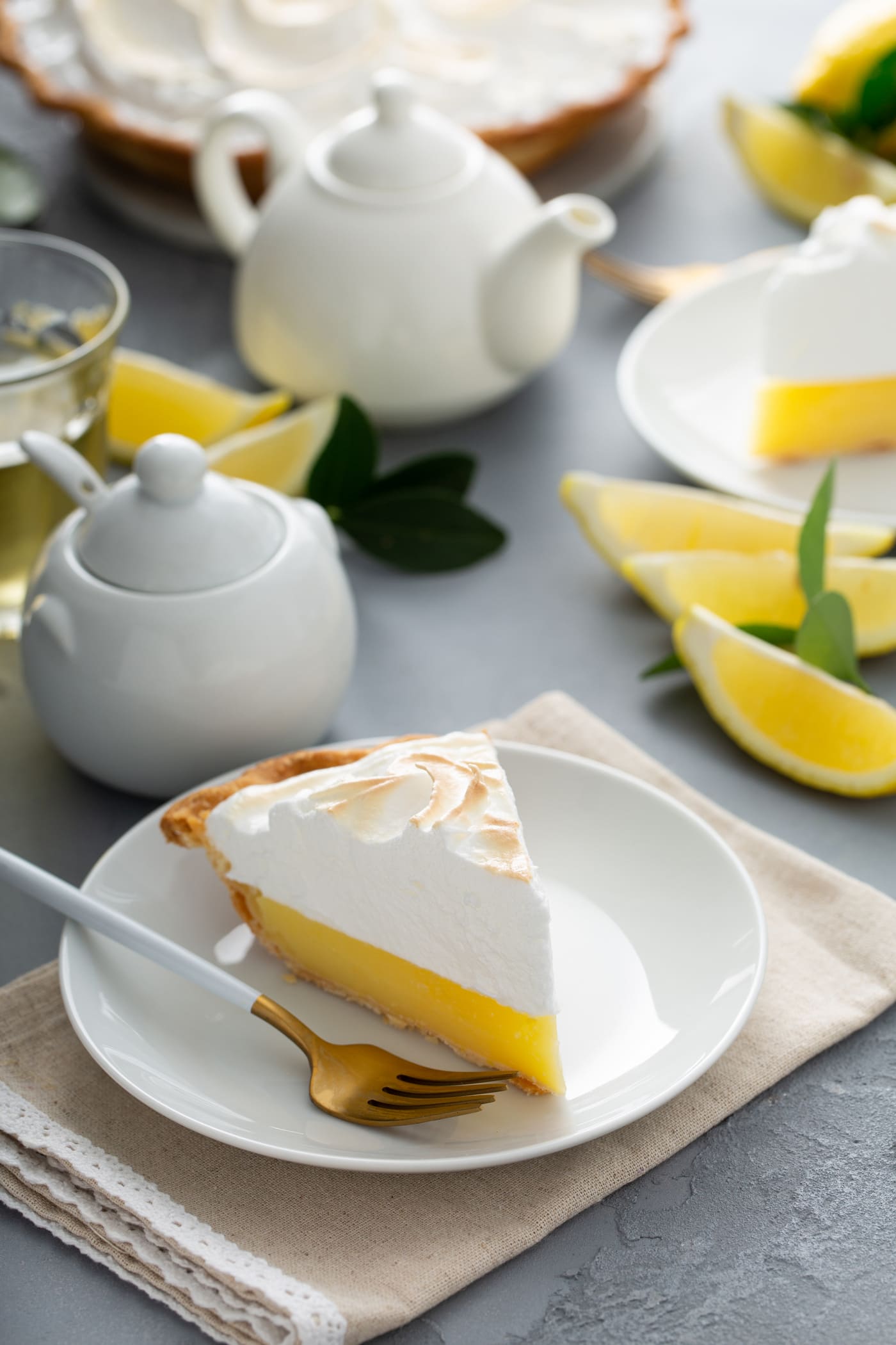 A piece of lemon meringue pie on a dessert plate. A fork in next to the pie and there are lemon wedges and a sugar bowl and cream pitcher in the background.