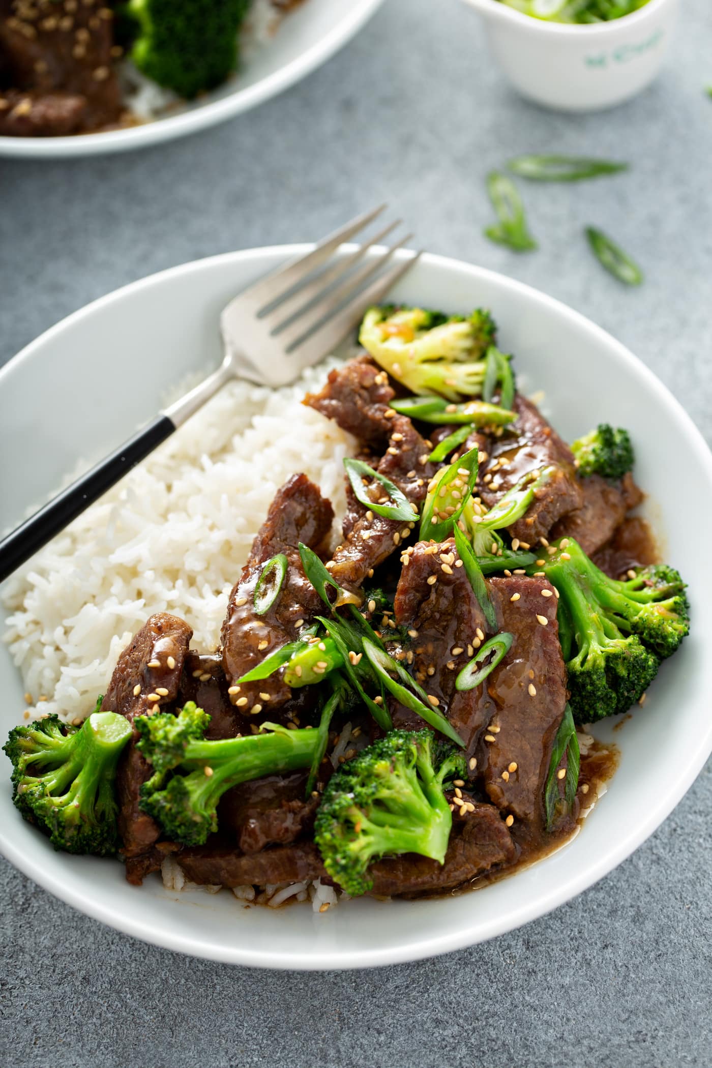 A photo of beef and broccoli on a white plate next to a serving of white rice and garnished with sliced green onions and toasted sesame seeds