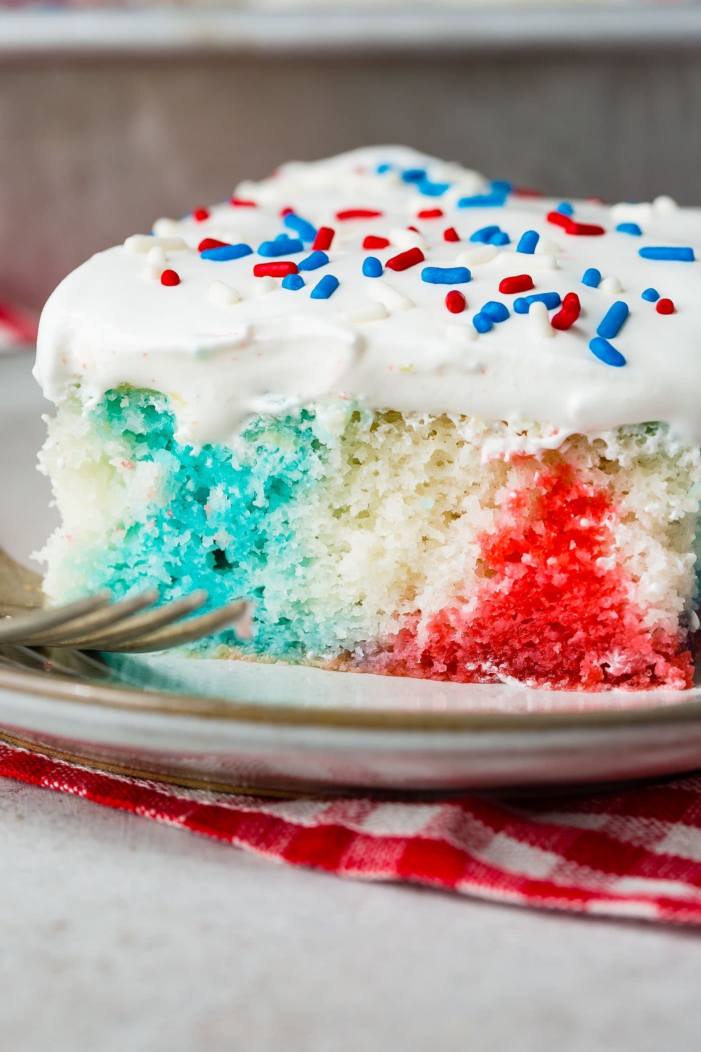 A piece of poke cake with a fork resting next to the cake. The cake is red, white and blue and is topped with white frosting and red, white and blue sprinkles. It is sitting on a red and white checked napkin.