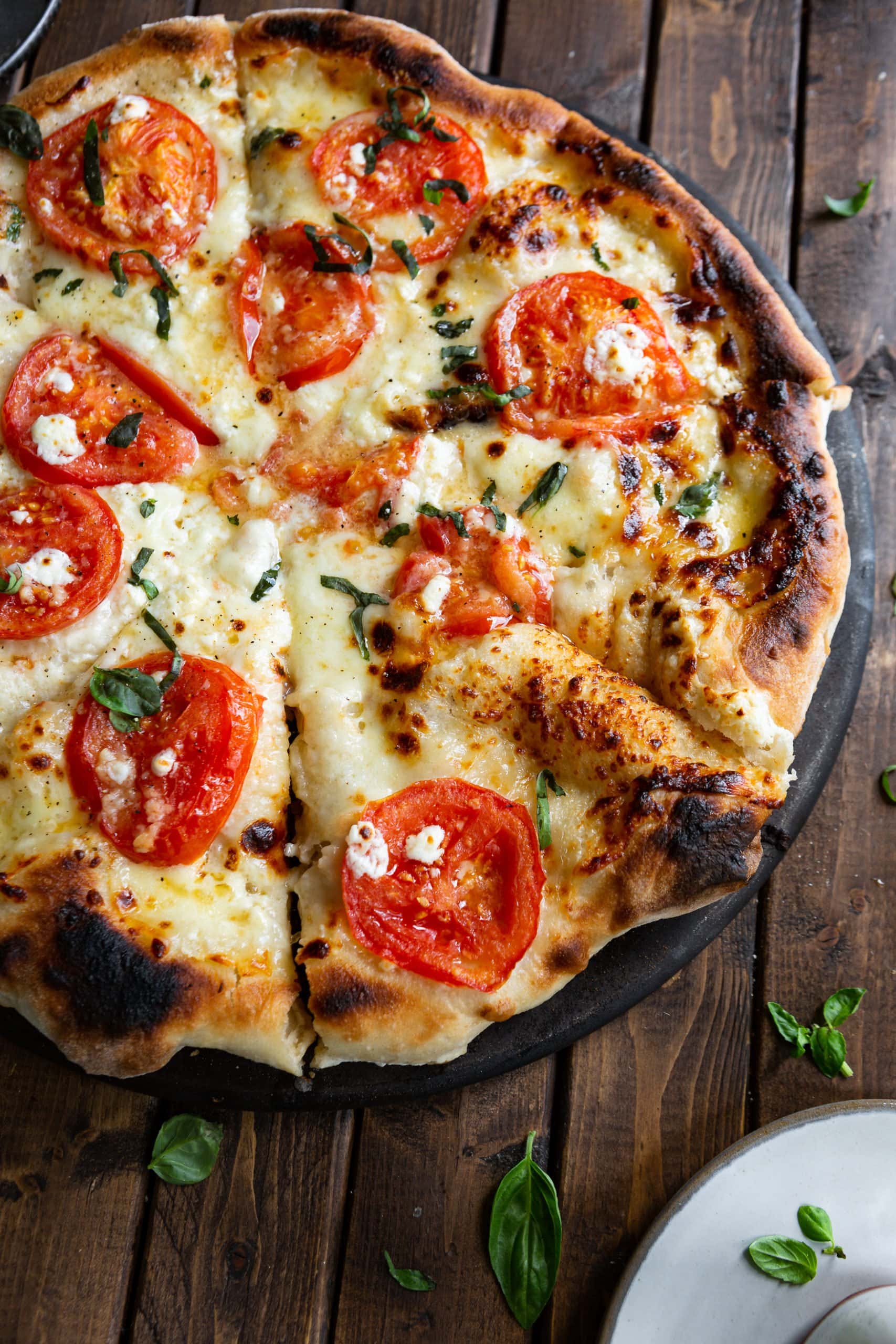 a photo of a whole margherita pizza that has been sliced into pieces topped with slices of tomato, chunks of feta cheese, chopped fresh basil and golden melted cheeses.