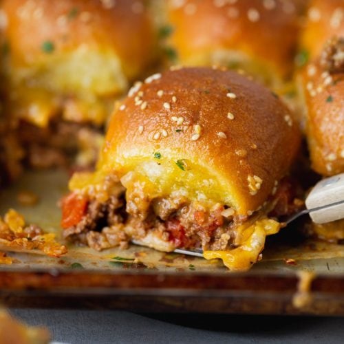 A cheeseburger slider filled with ground beef, melty cheese and chopped tomatoes. there are other sliders in the background.