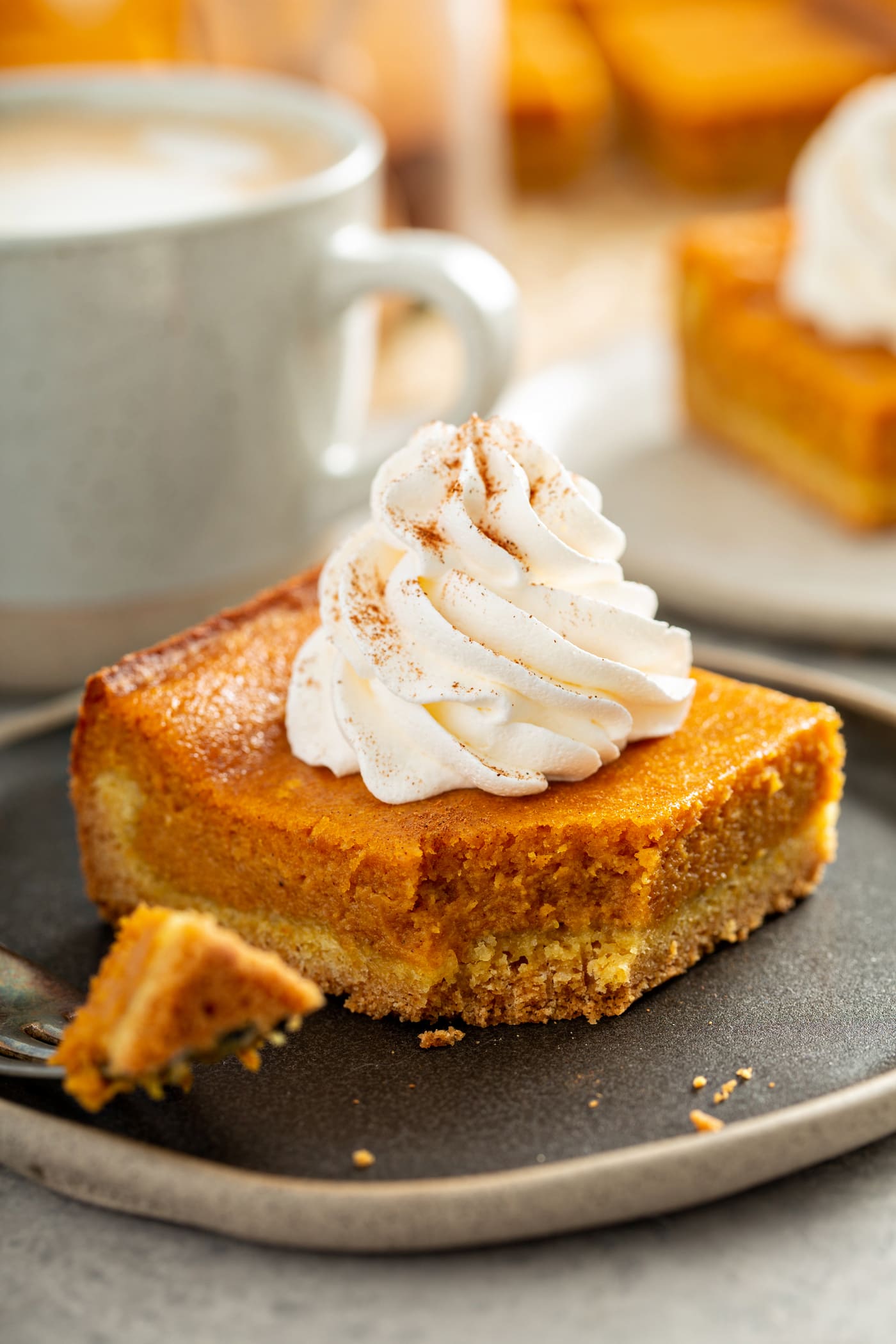 A square piece of pumpkin gooey butter cake with a swirl of whipped cream on top. A fork with a bite of cake is laying on the plate and a white cup and white plate with a piece of pumpkin cake are in the background. 