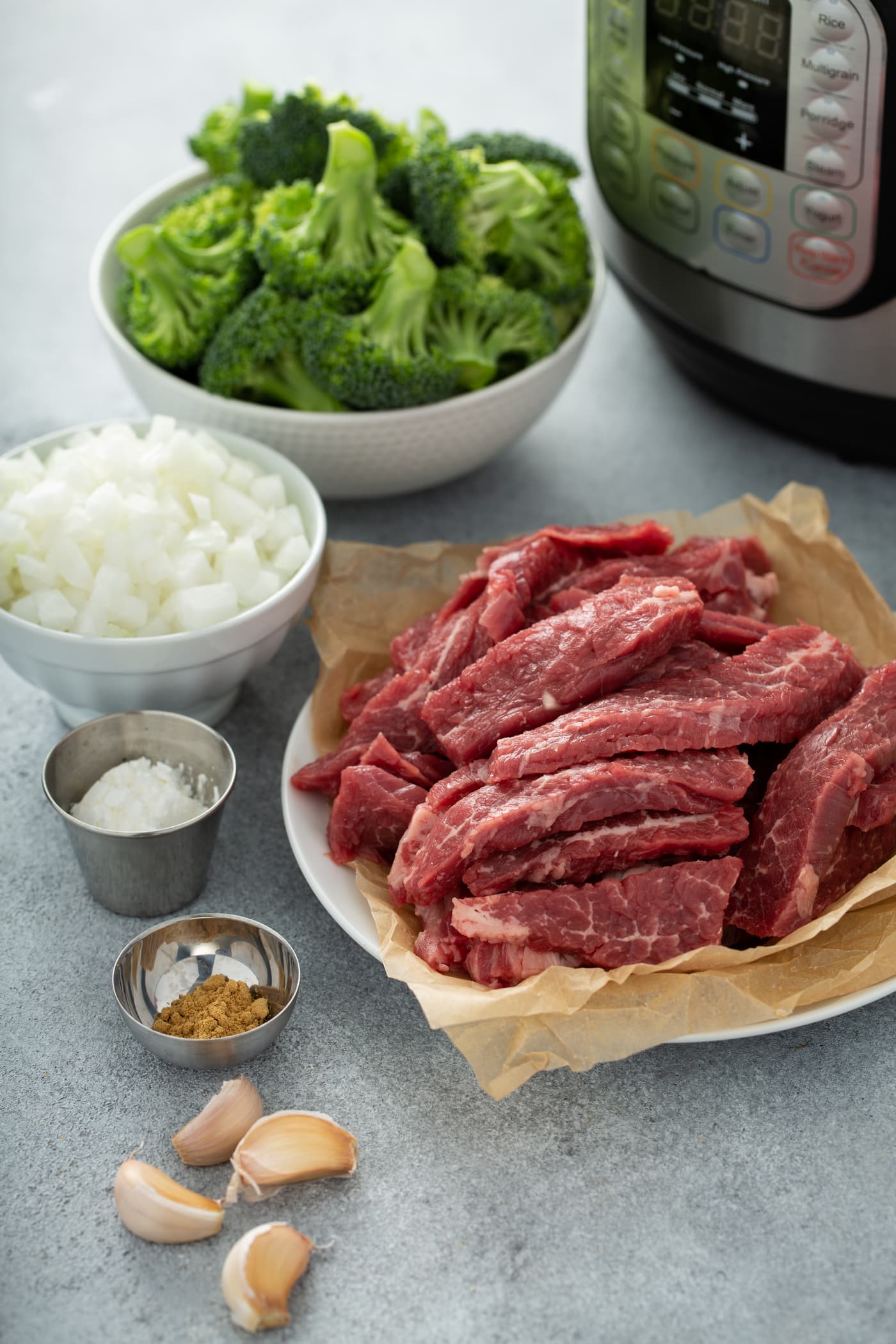 A photo of the ingredients needed to make beef and broccoli with an Instant Pot in the background. The ingredients are in separate bowls, including broccoli florets, chopped onion, sliced raw flank steak, cornstarch, ground ginger and garlic cloves.