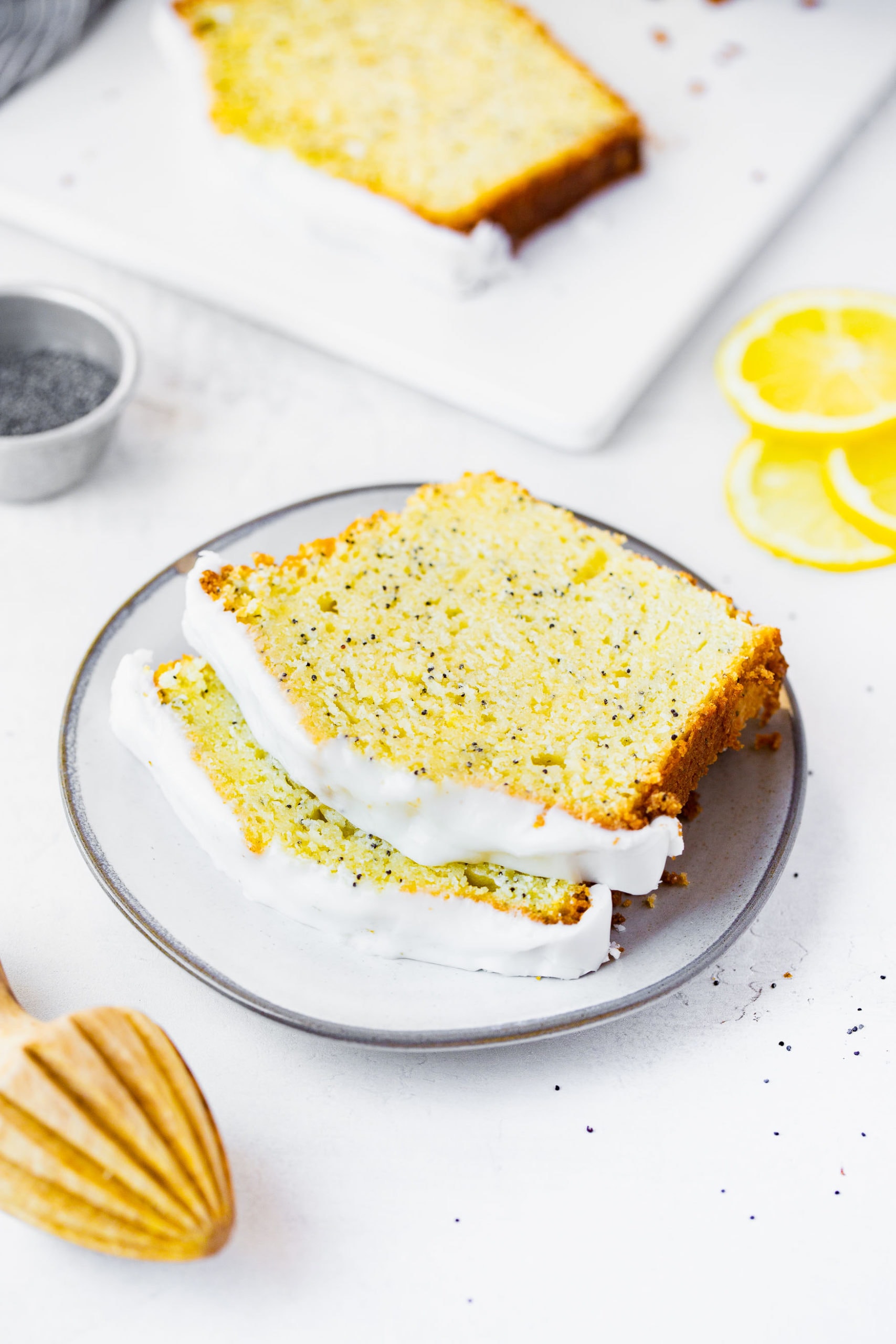 a photo of two slices of lemon poppy seed loaf cake stacked on each other on a small white plate with lemon slices in the background and a wooden lemon reamer in the foreground.