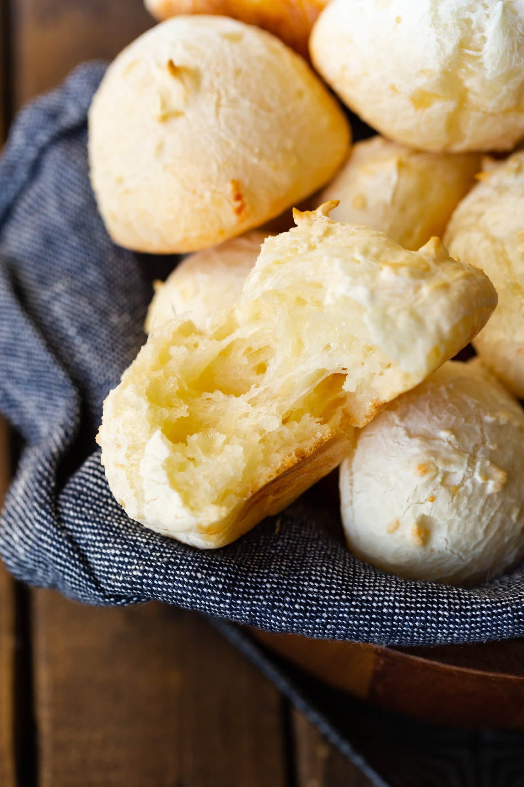 a photo of a piece of pao de queijo that has been torn in half so you can see the cheesy middle sitting on top of a pile of more golden cheese rolls.