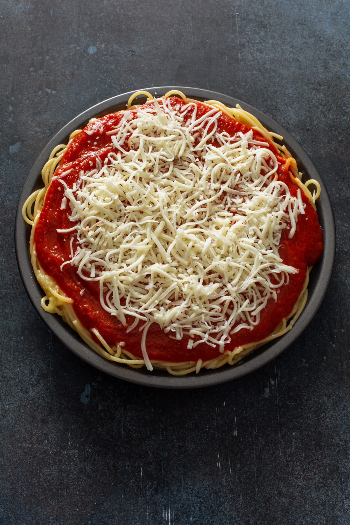 A pie dish filled with cooked spaghetti, topped with spaghetti sauce and shredded cheeses.