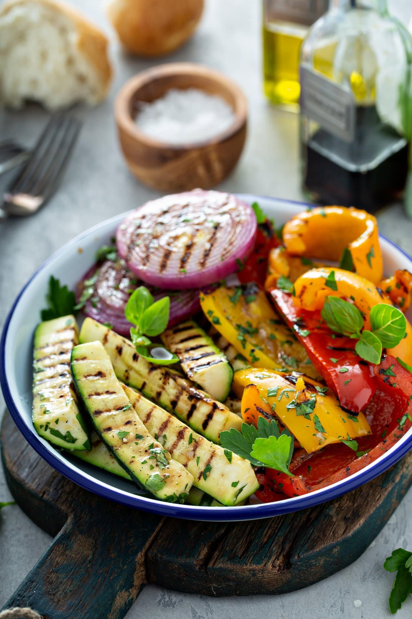 A plate of grilled vegetables. The zucchini, bell peppers, and red onions have beautiful grill marks and a few parsley leaves are scattered on top. There is a small wooden bowl of salt, a fork, and a glass cup of balsamic glaze in the background.