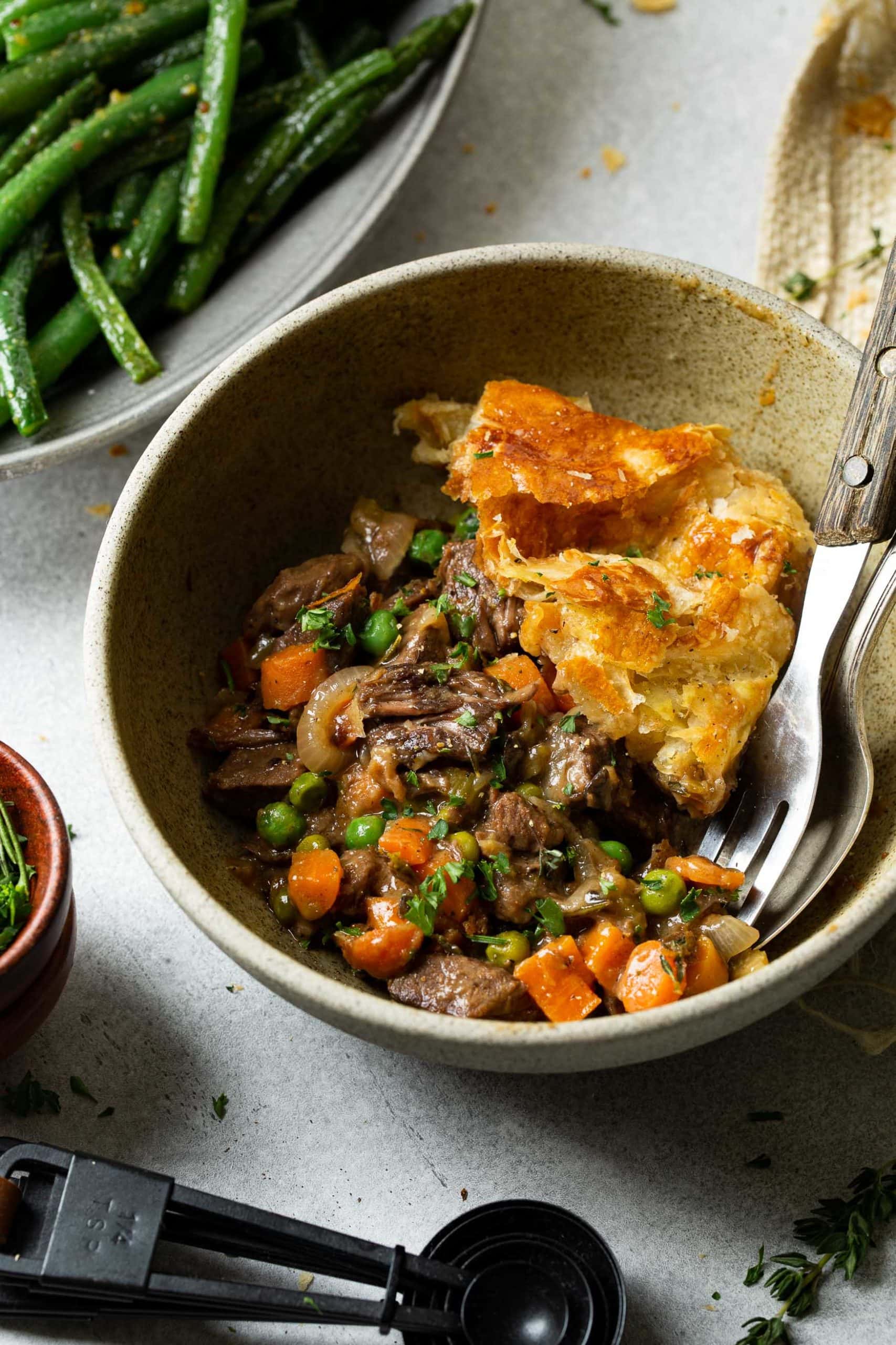 A photo of a serving of beef pot pie in a bowl with chunks of beef, carrots, peas, caramelized onions all in a gravy topped with a golden puff pastry.